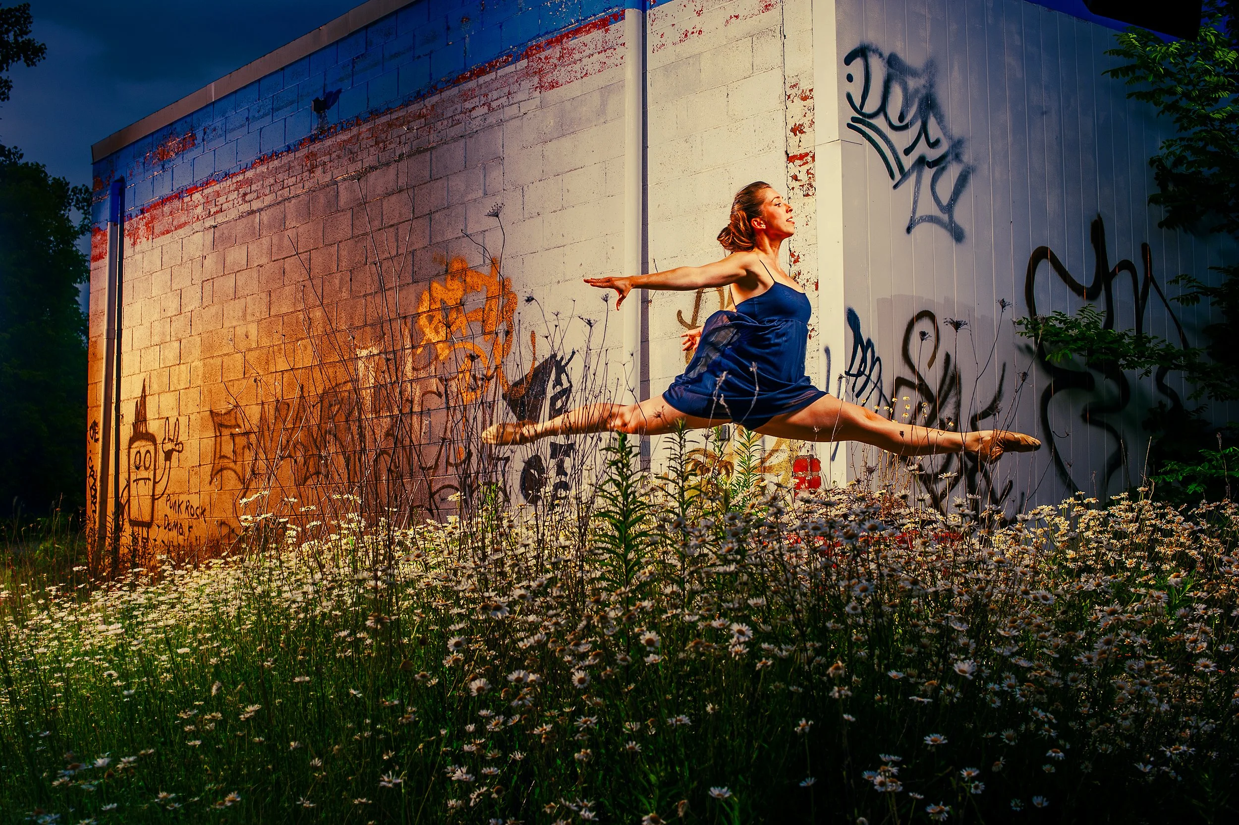 A woman in a blue dress performing a ballet stretch outdoors at dusk, with graffiti-covered walls and wildflowers in the foreground.