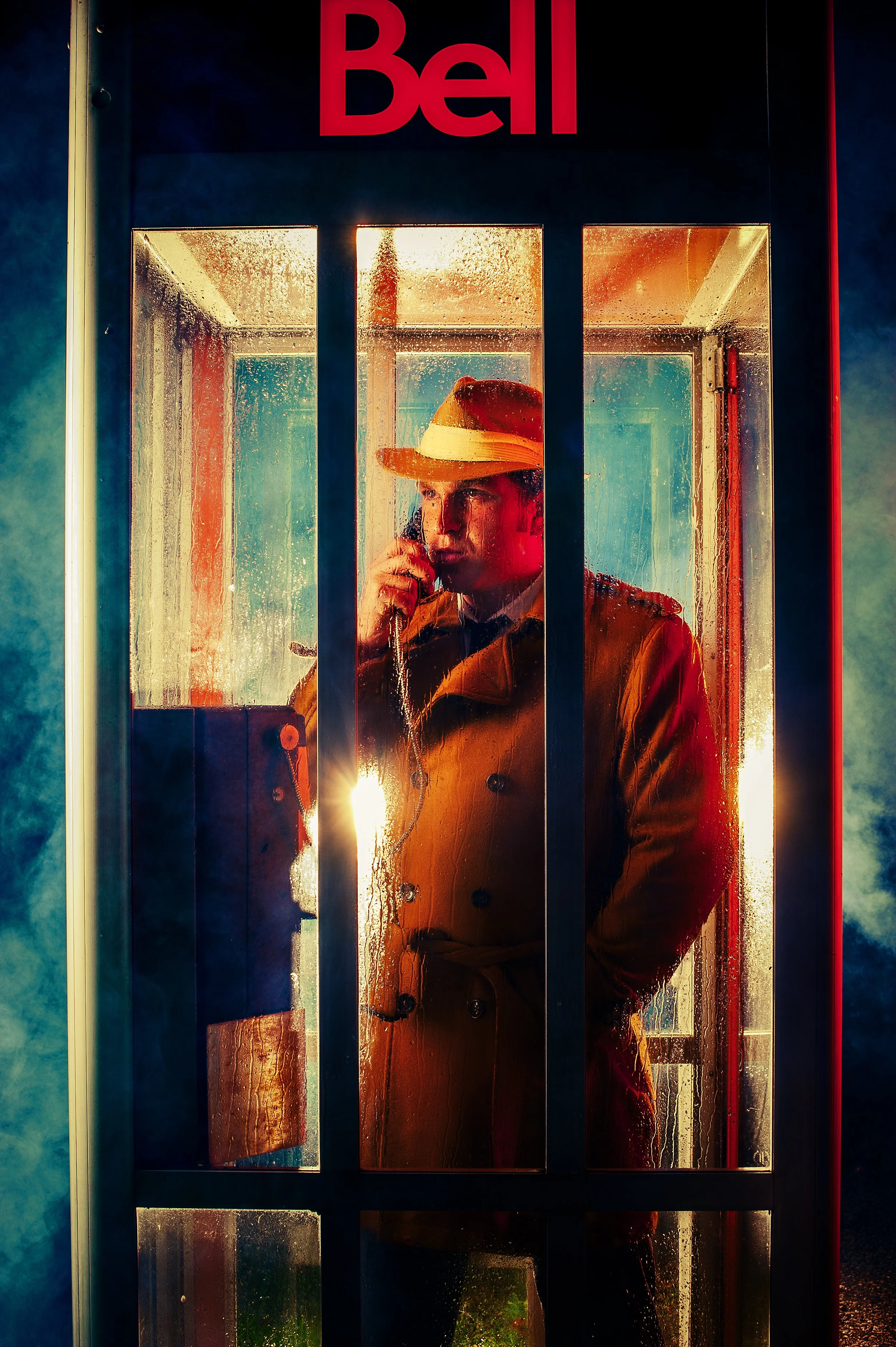 A woman with dark hair, wearing a beige hat and beige trench coat, talking on a phone inside a rainy phone booth at sunset or nighttime, with raindrops on the glass and a red Bell sign above.