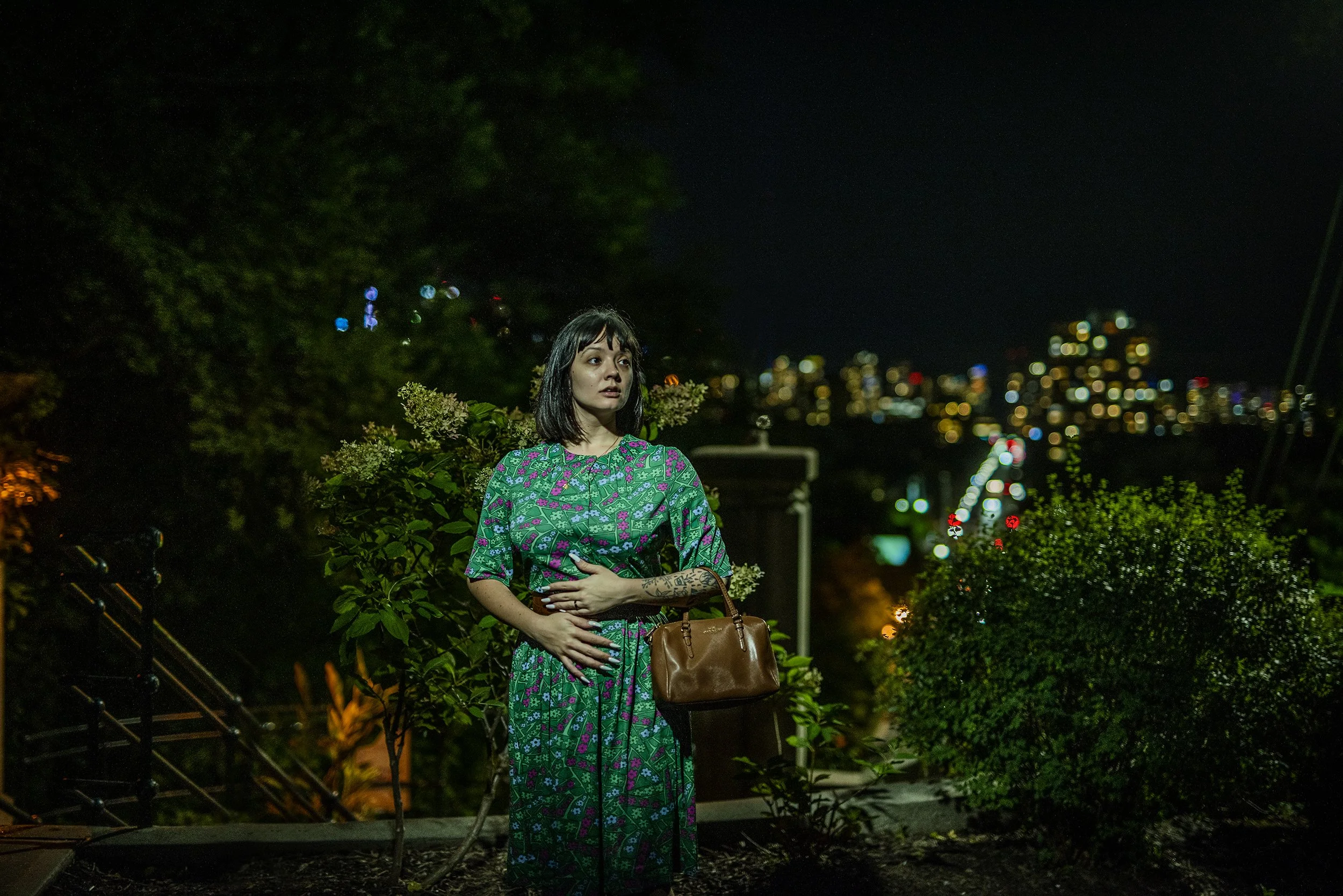 A young woman with dark hair, wearing a patterned dress, stands outdoors at night with city lights in the background. She is holding a brown handbag and has tattoos on her arm, surrounded by greenery.