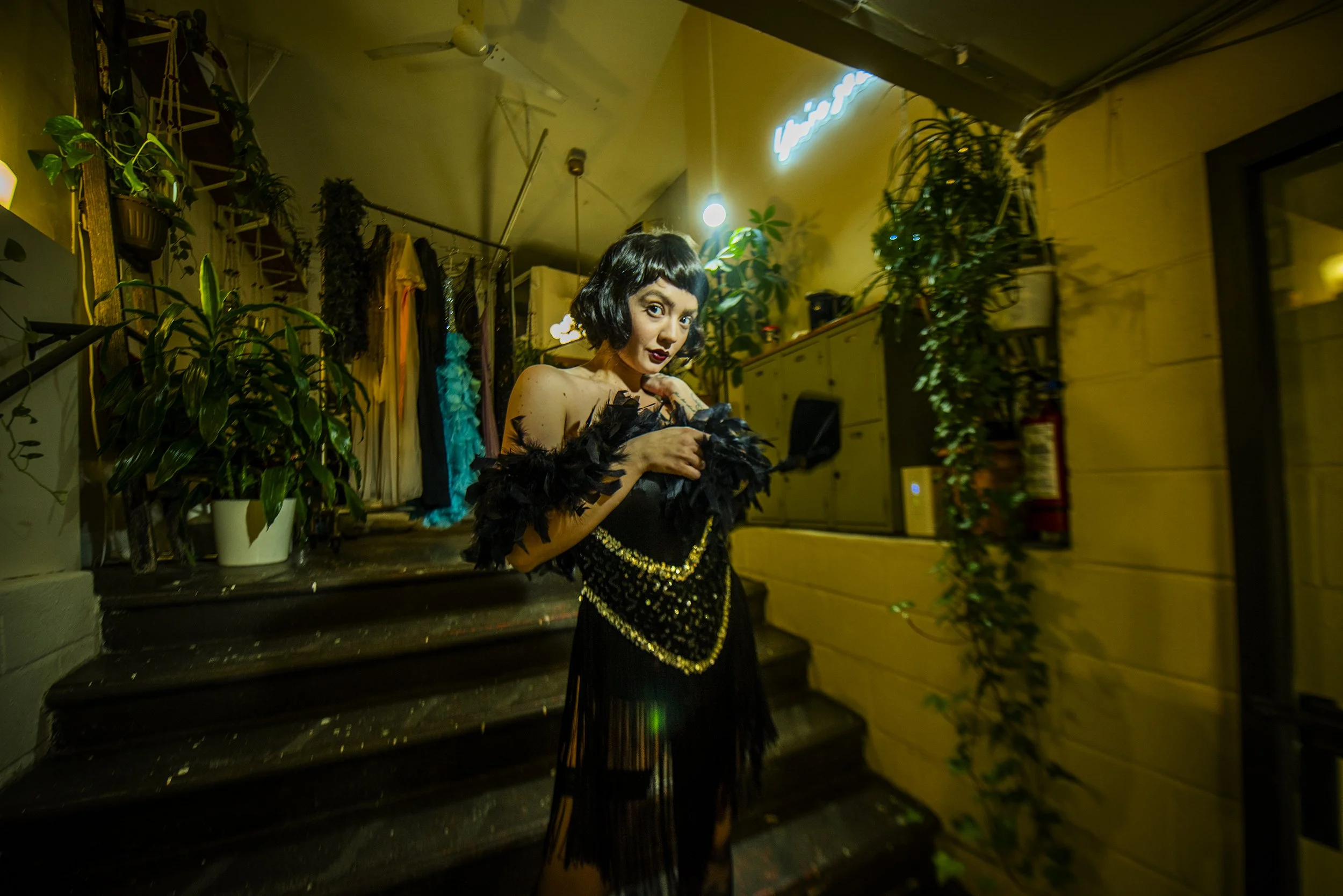 A woman with short black hair in a wig is standing inside a room decorated with green plants, yellow walls, and clothing hanging in the background. She is wearing a black dress with gold embellishments and feathered accessories, posing with a hand ne