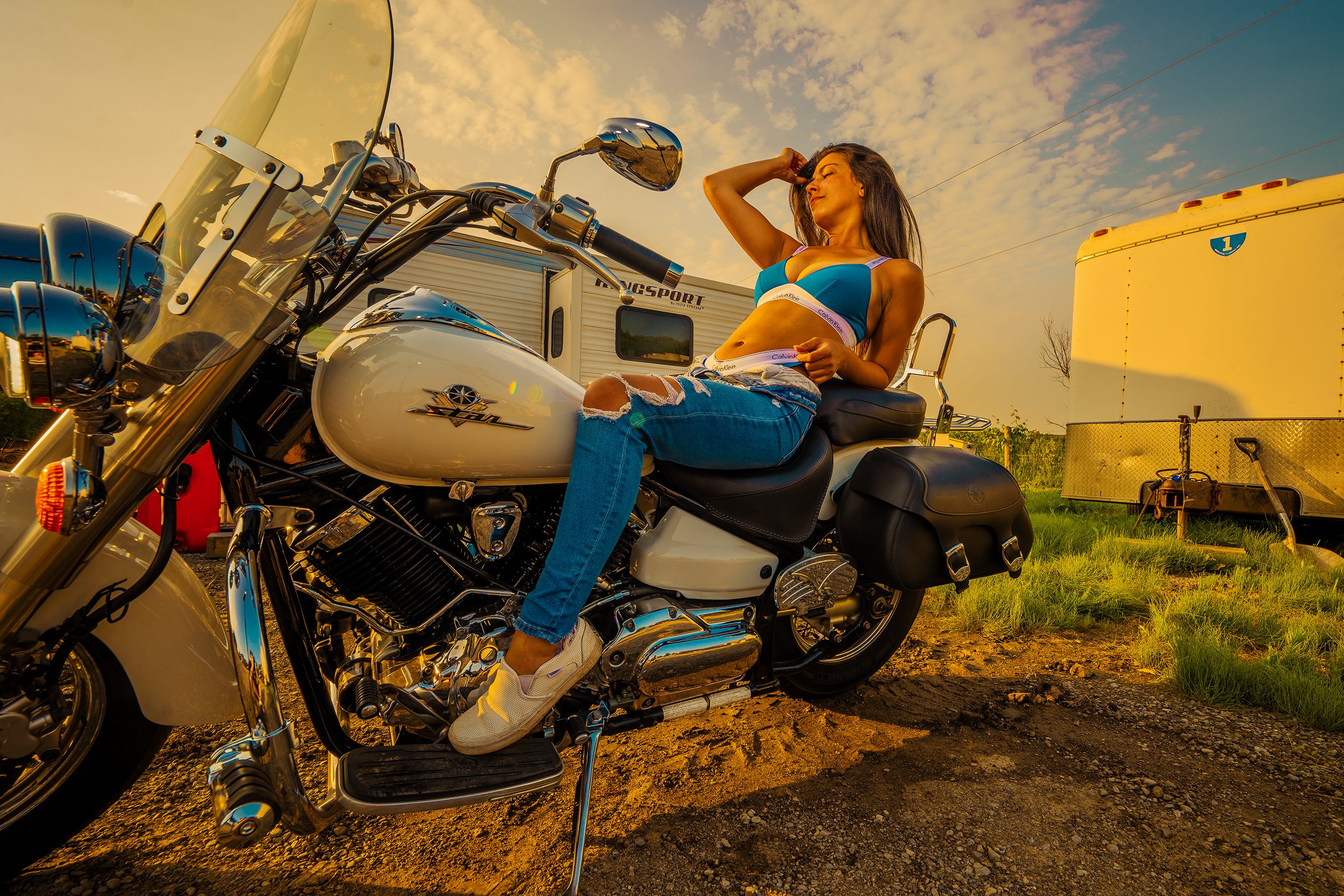A woman sitting on a white motorcycle during a sunset, wearing a blue sports bra, ripped jeans, and sneakers, with trailers and grassy terrain in the background.