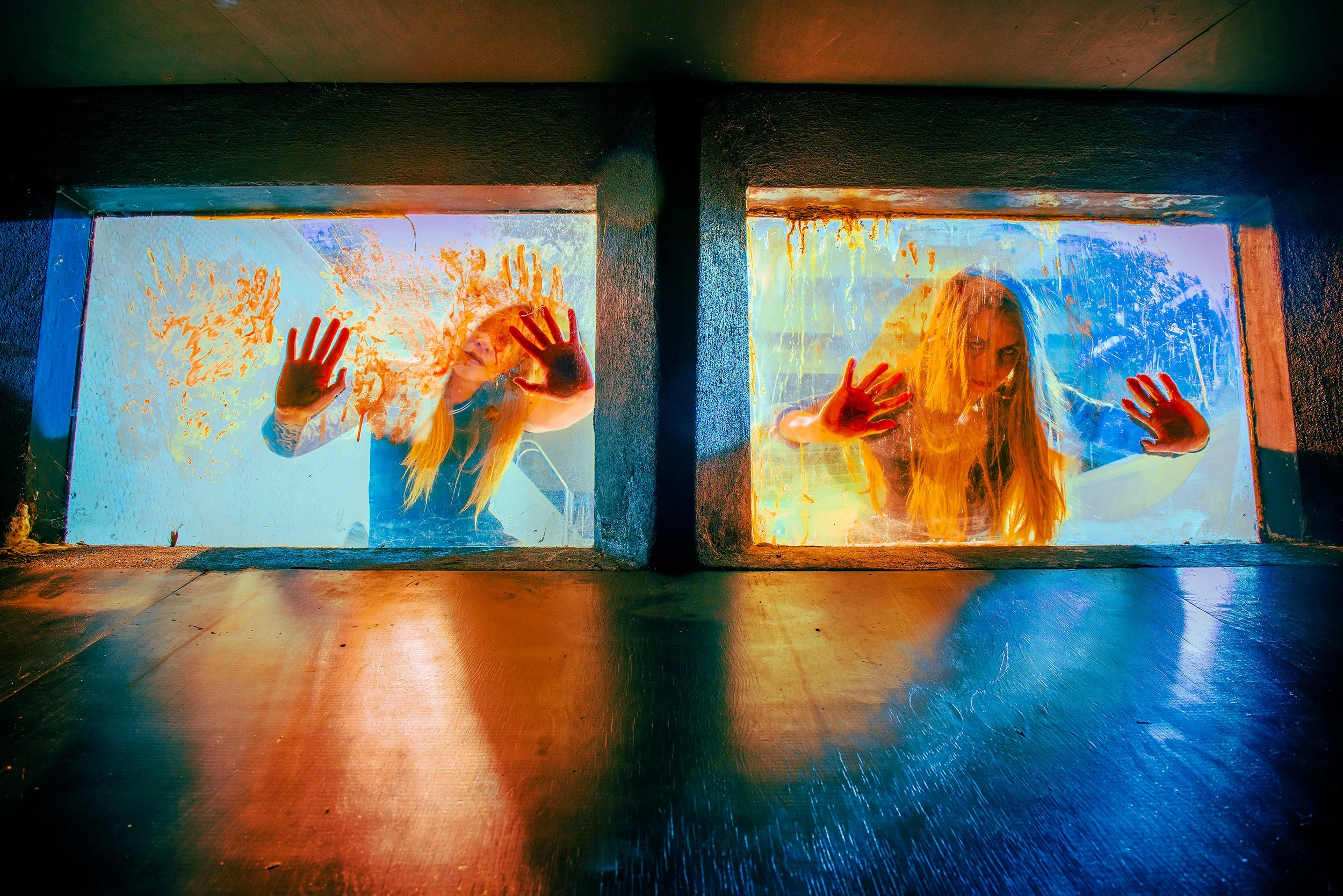 Two women with painted faces pressed against glass windows, with colorful lighting.