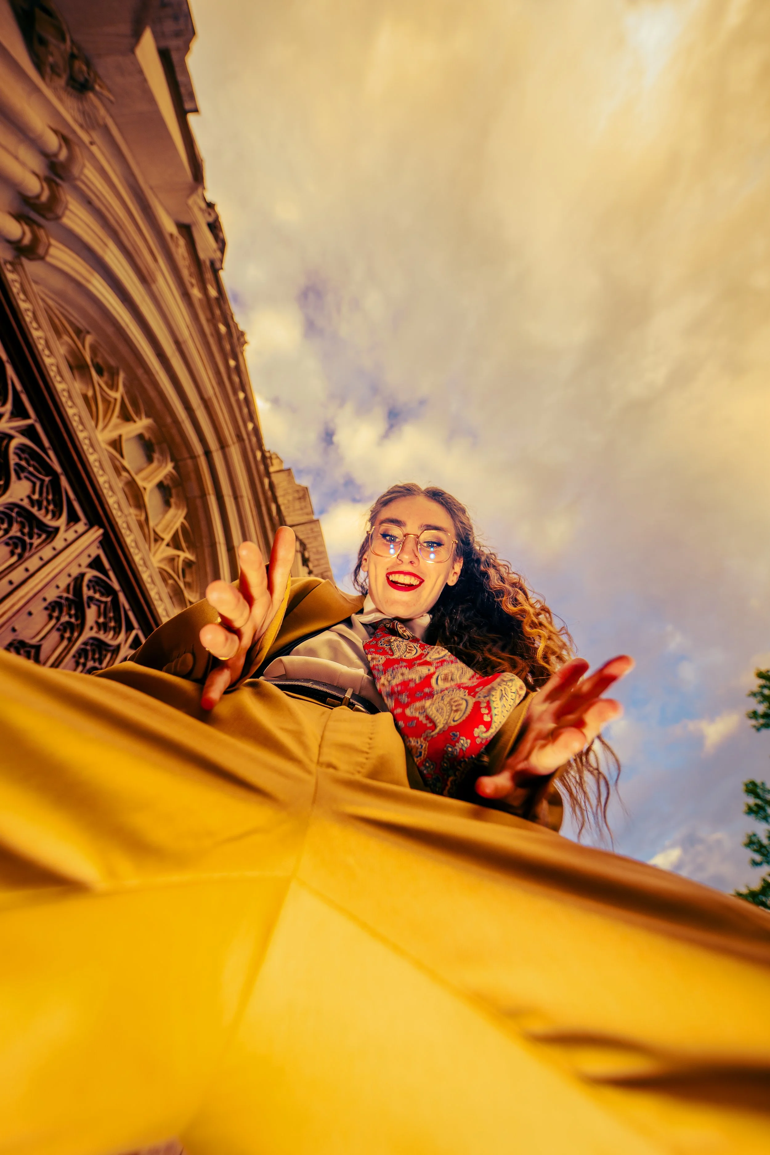 A young woman with curly hair, wearing glasses, a patterned scarf, and a yellow jacket, smiling and making peace signs with her hands, standing outdoors near an ornate building with a decorative balcony railing under a cloudy sky.