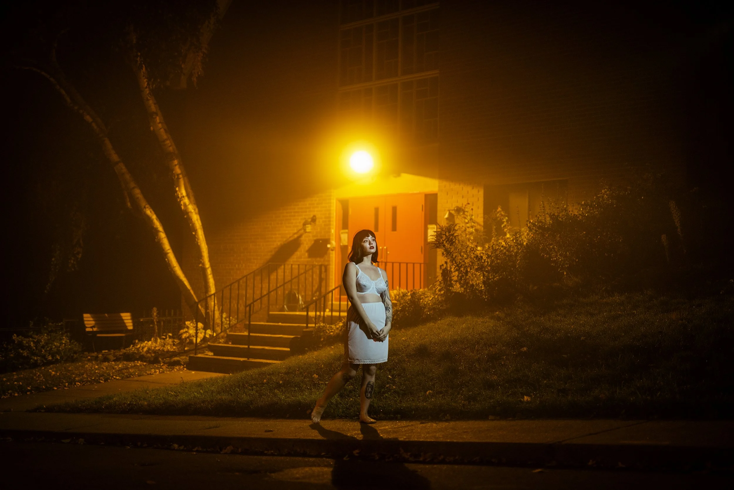 A woman with dark hair and tattoos on her legs standing outside at night in front of a house with bright orange lighting, wearing a white lace dress.