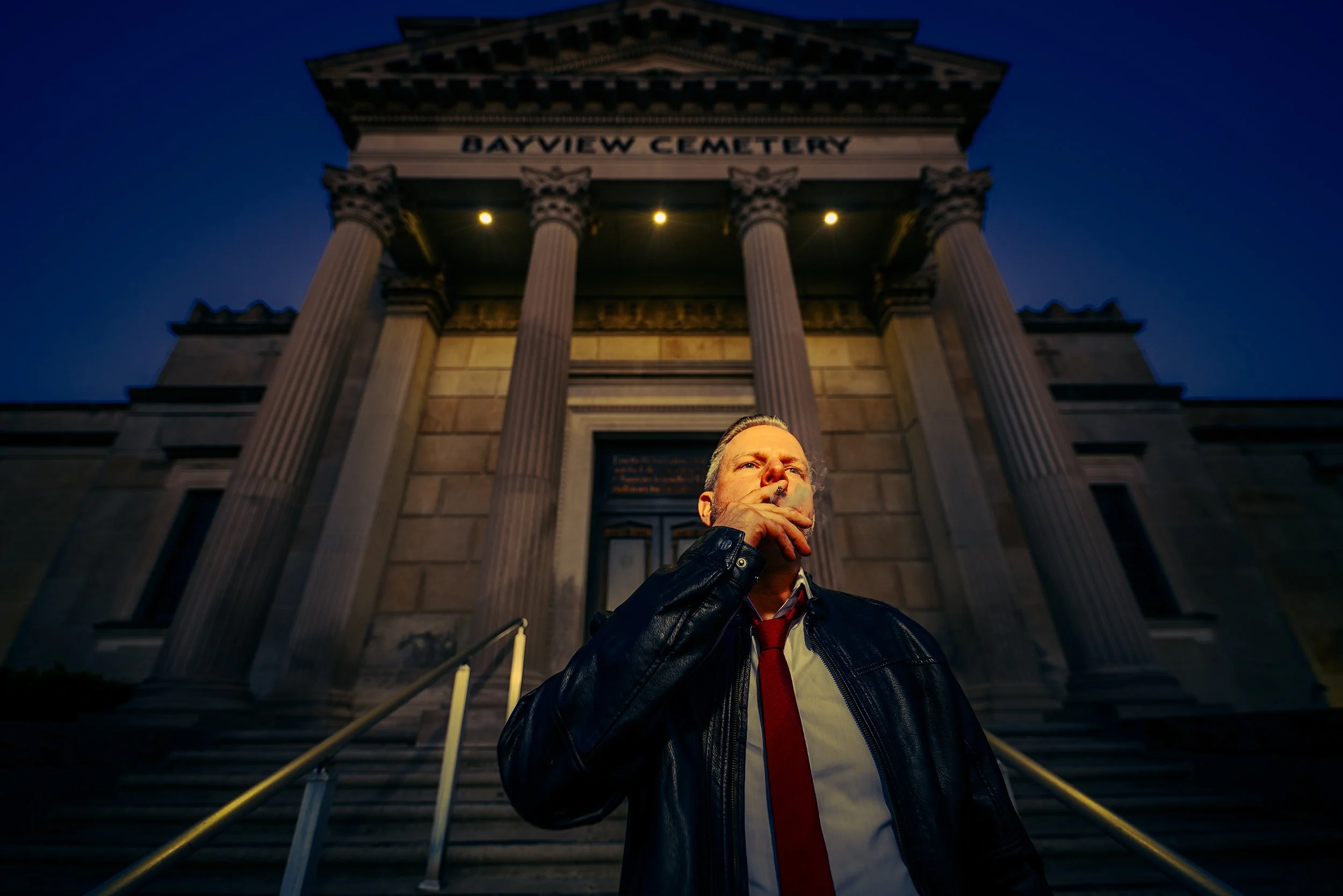 A man in a leather jacket and red tie smoking a cigarette in front of a historic building with large columns labeled "Bayview Cemetery" at dusk.