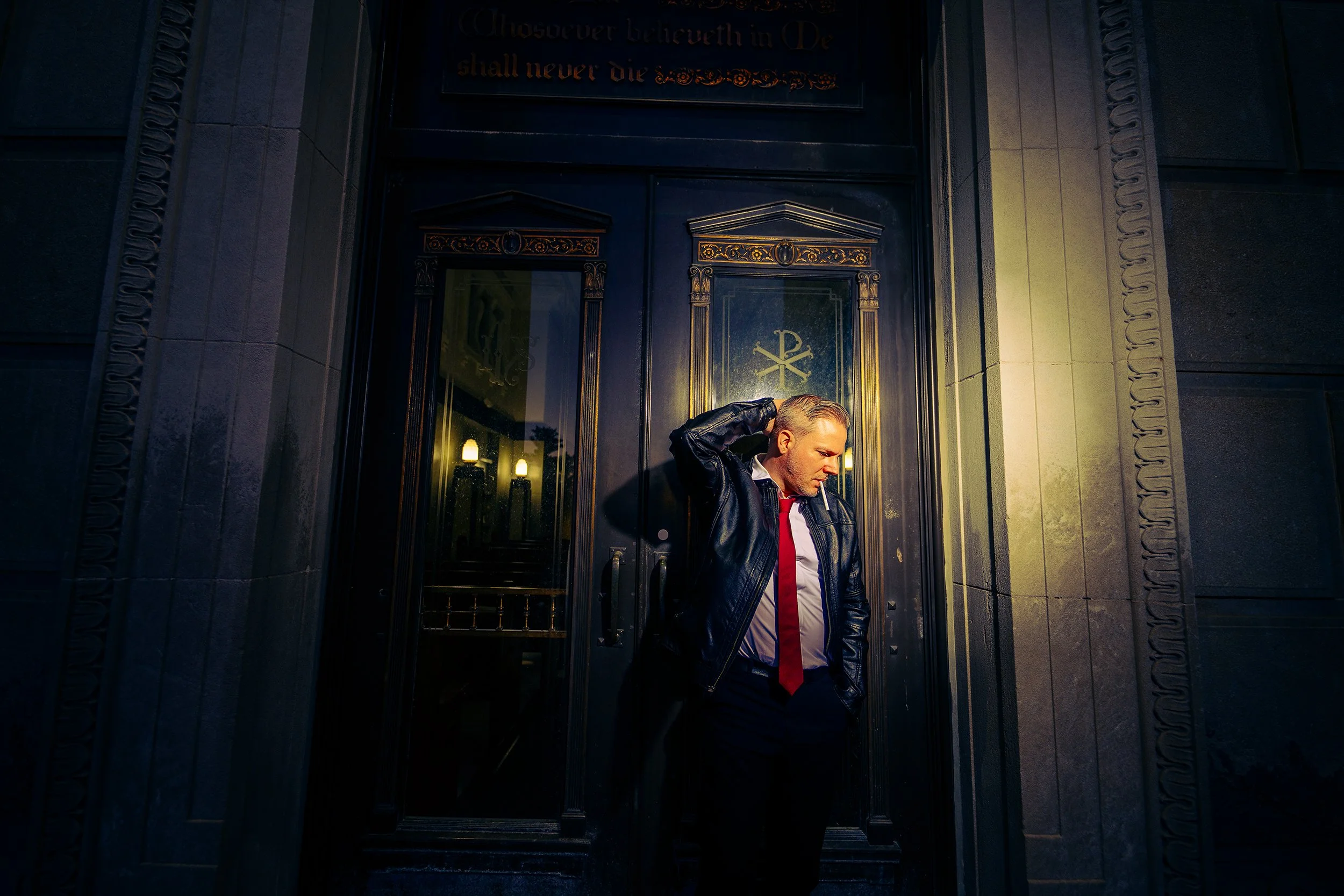 A man in a leather jacket, white shirt, and red tie standing outside a dark building with ornate doors. He is holding his head with one hand and smoking a cigarette.