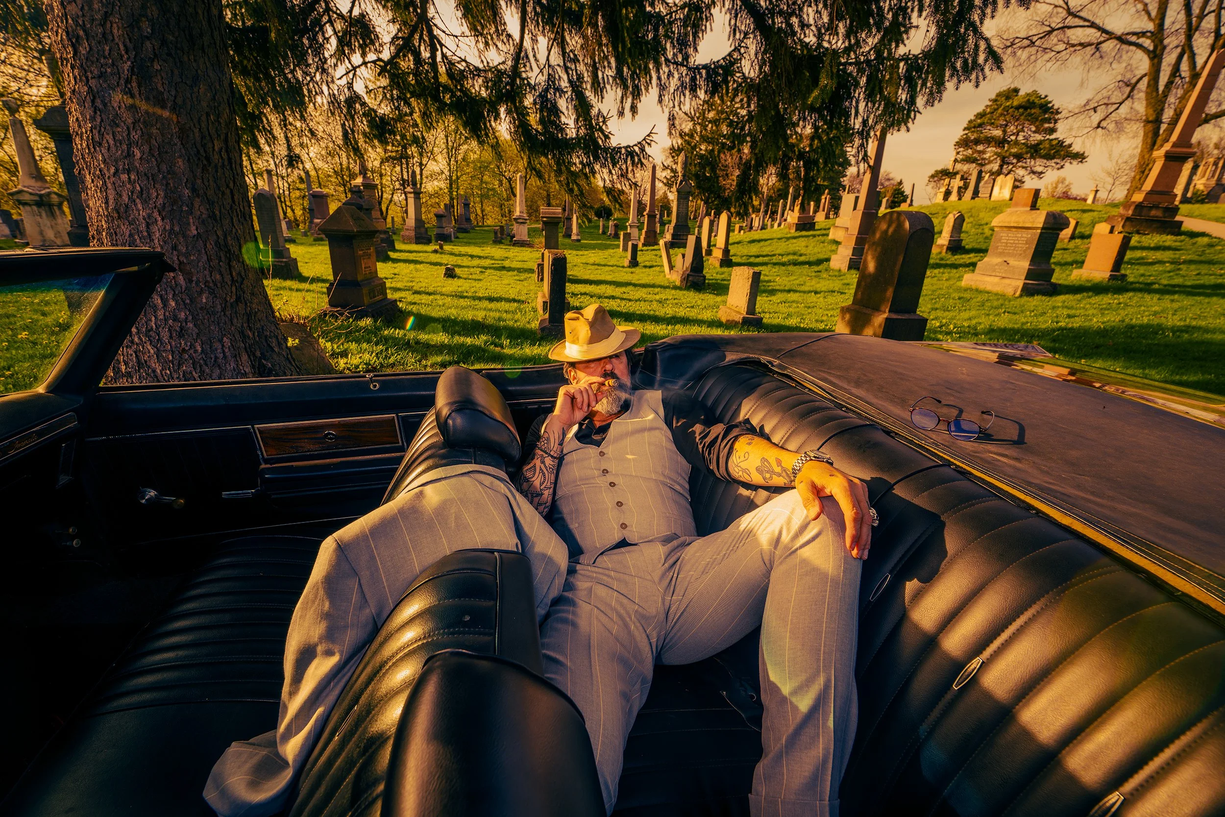 A man with sunglasses, tattoos, and a hat reclining inside a black vintage car, smoking a cigar, with a graveyard in the background during golden hour sunset.