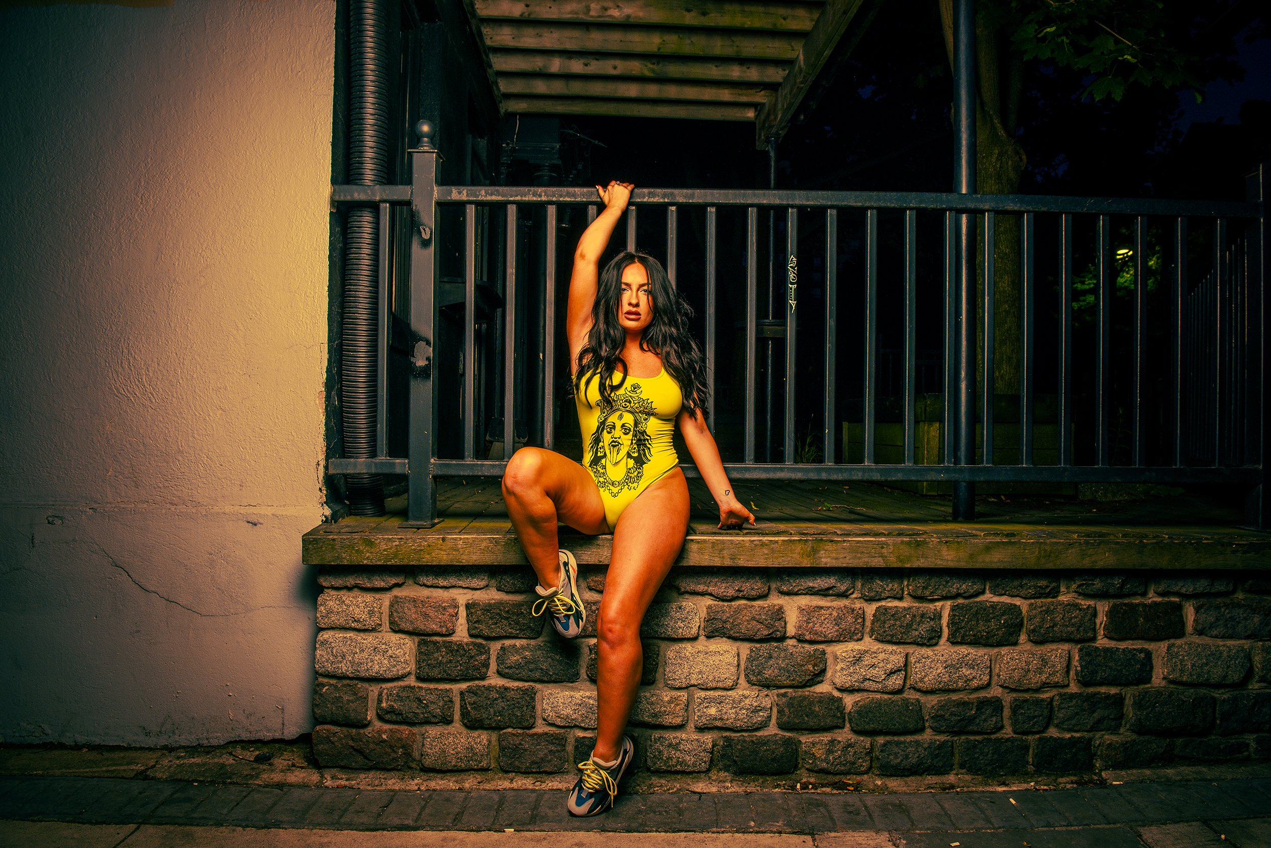 A woman in a yellow swimsuit with a graphic design sitting on a brick ledge in front of a black metal gate at night.