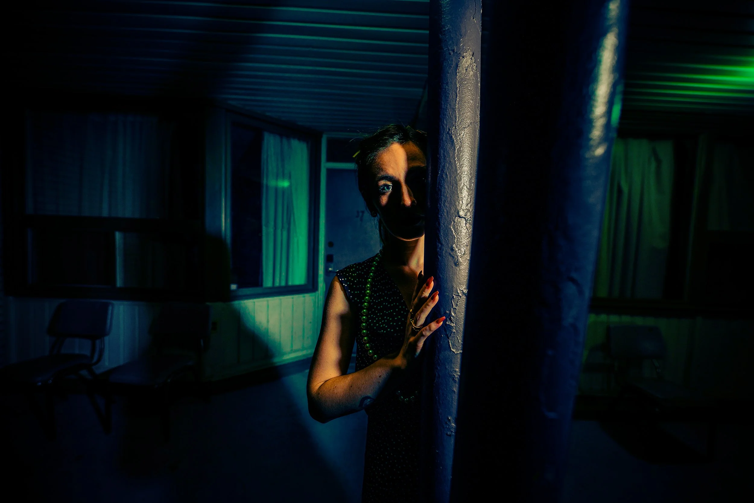 A woman peeking from behind a pillar in a dimly lit room, illuminated by neon green and blue lights, wearing a black dress with white polka dots and a green beaded necklace.