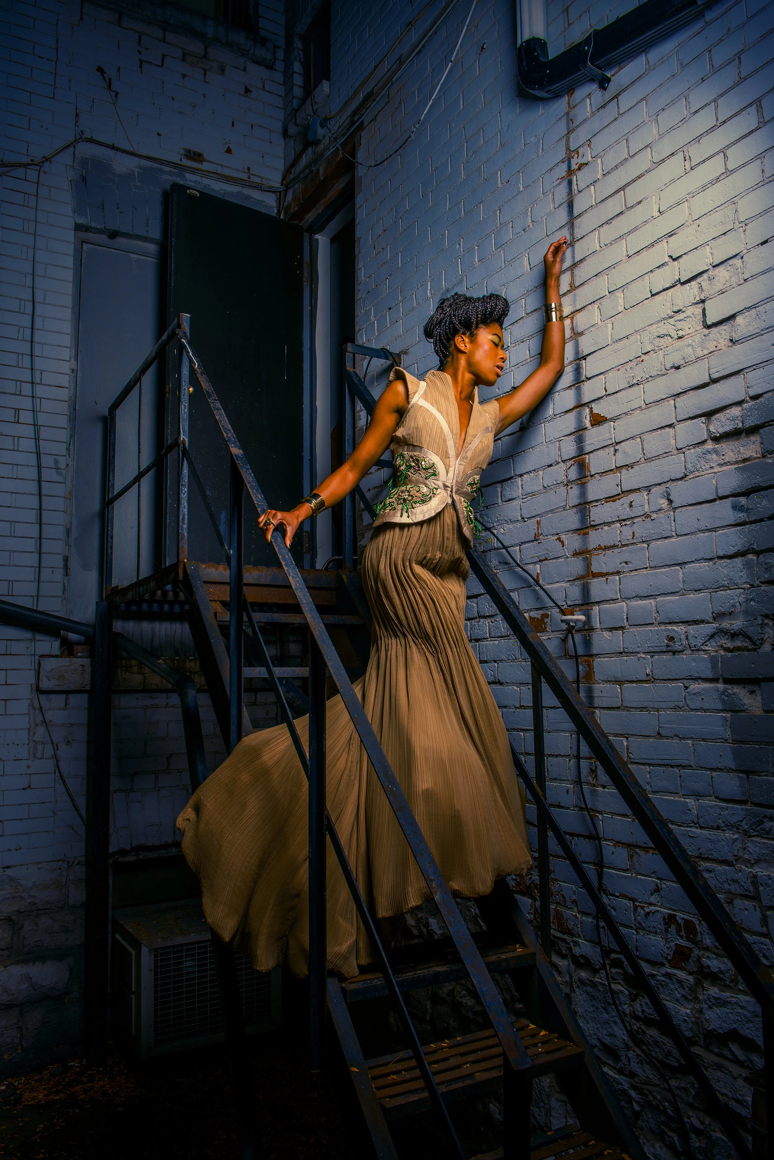A woman in a beige and green dress poses on an outdoor metal staircase against a gray brick wall, with her right arm raised and her eyes closed.