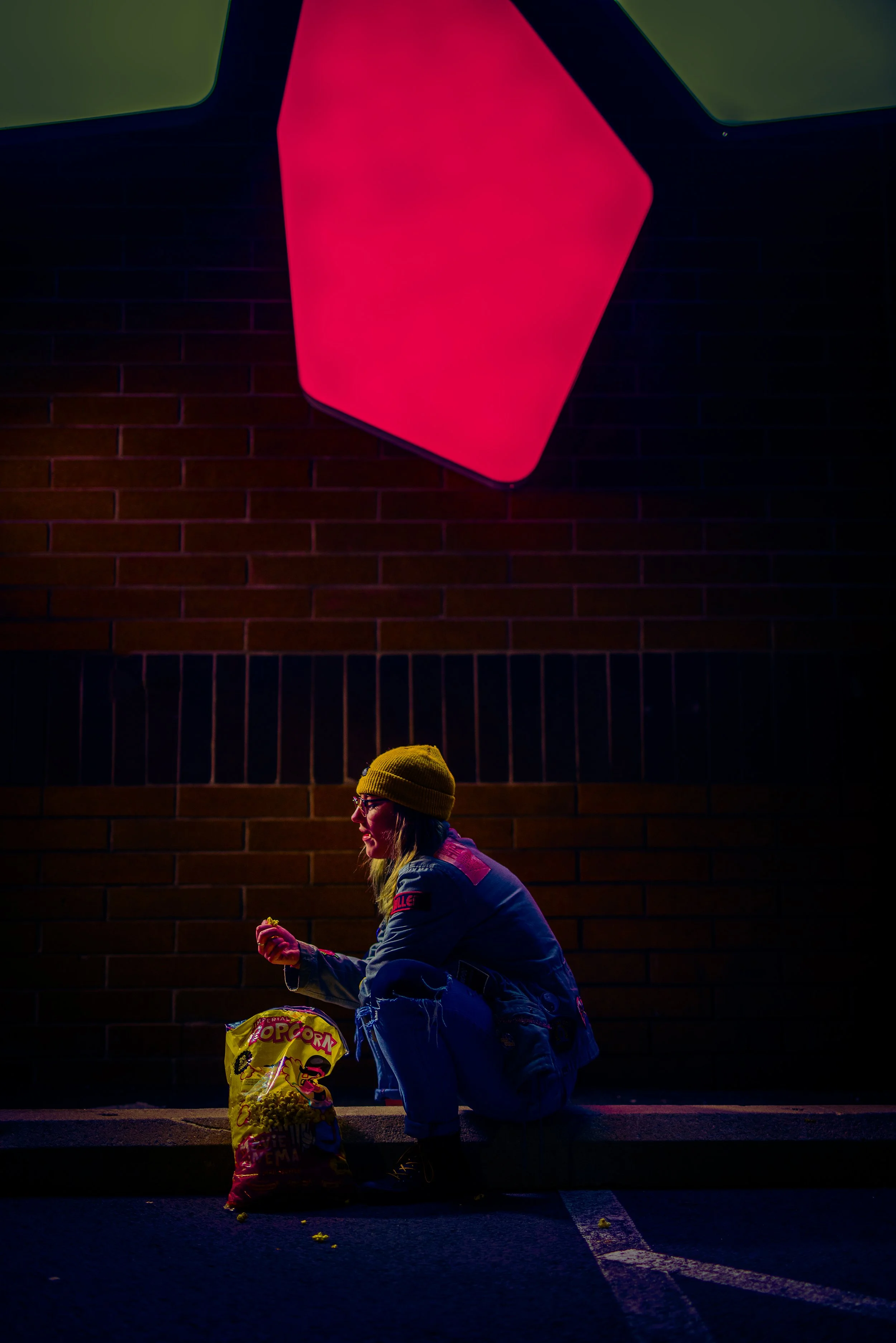 A woman in a yellow beanie and jean jacket eating popcorn while sitting on a curb at night. A large bag of popcorn is on the ground beside her, and she is holding a piece of popcorn in her hand. The scene is lit with colorful neon lights reflected on