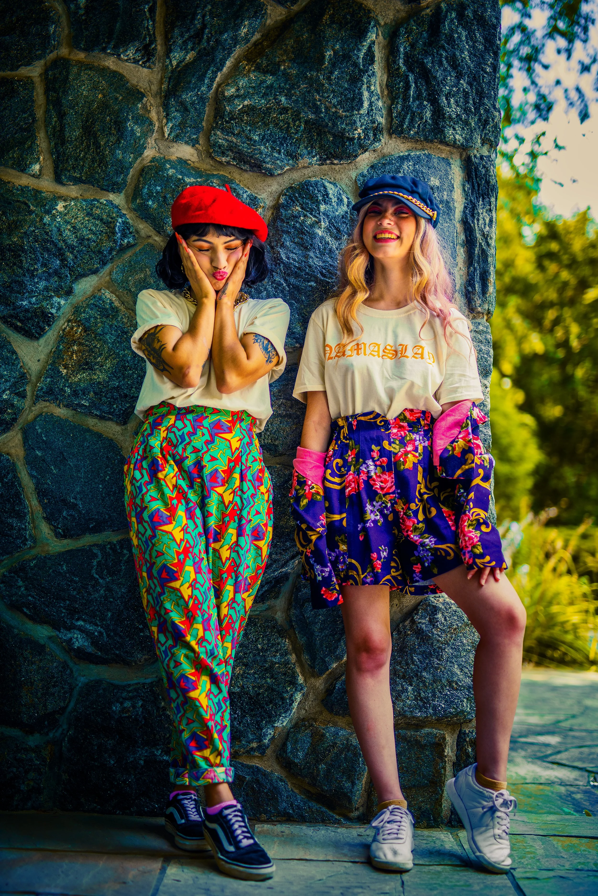 Two young women standing by a stone wall outdoors, smiling and laughing, dressed in colorful, casual festival-style clothing and hats, with trees in the background.