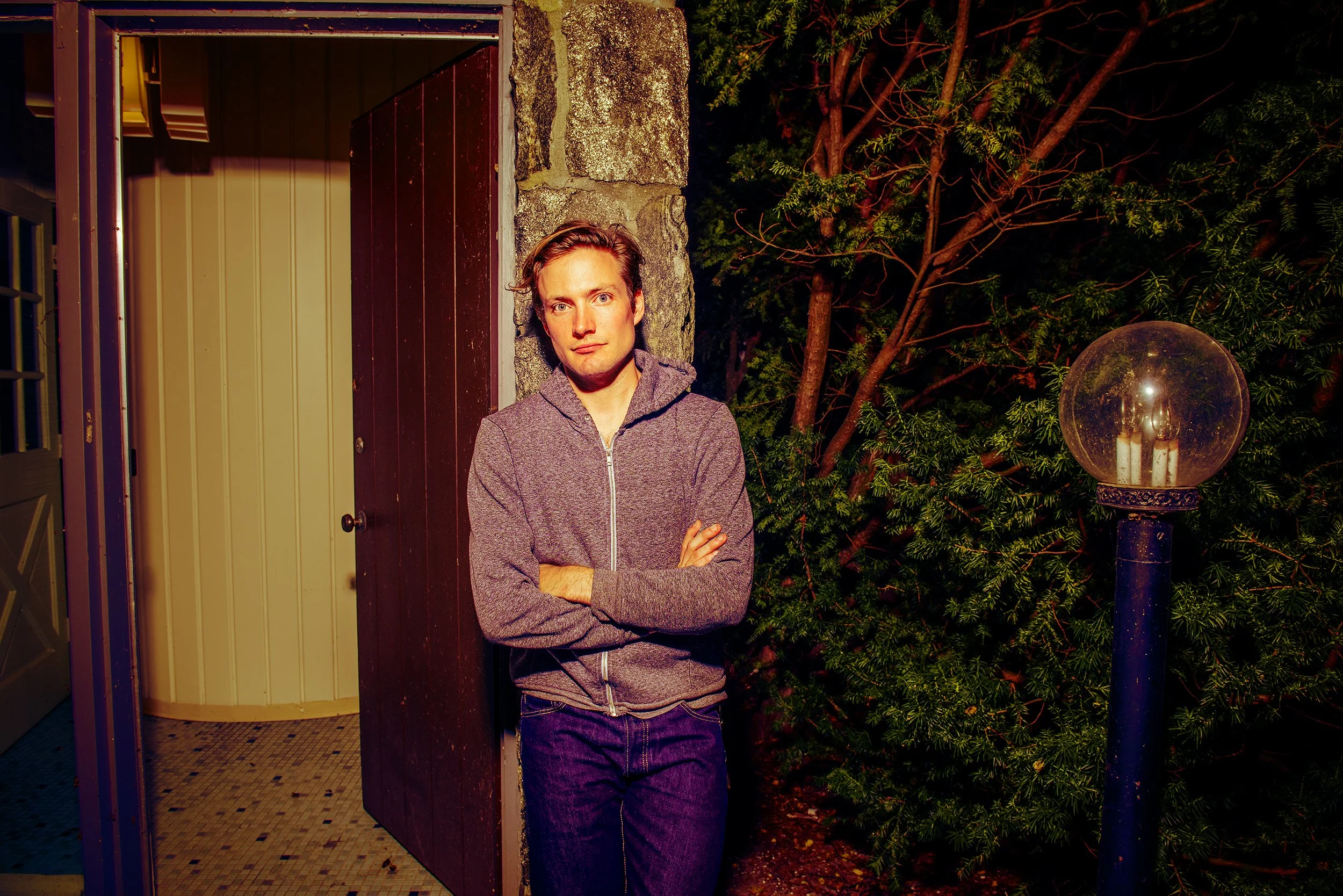 A man with crossed arms leaning against a stone wall near an open door, standing next to a tree and a garden lamp, during nighttime.