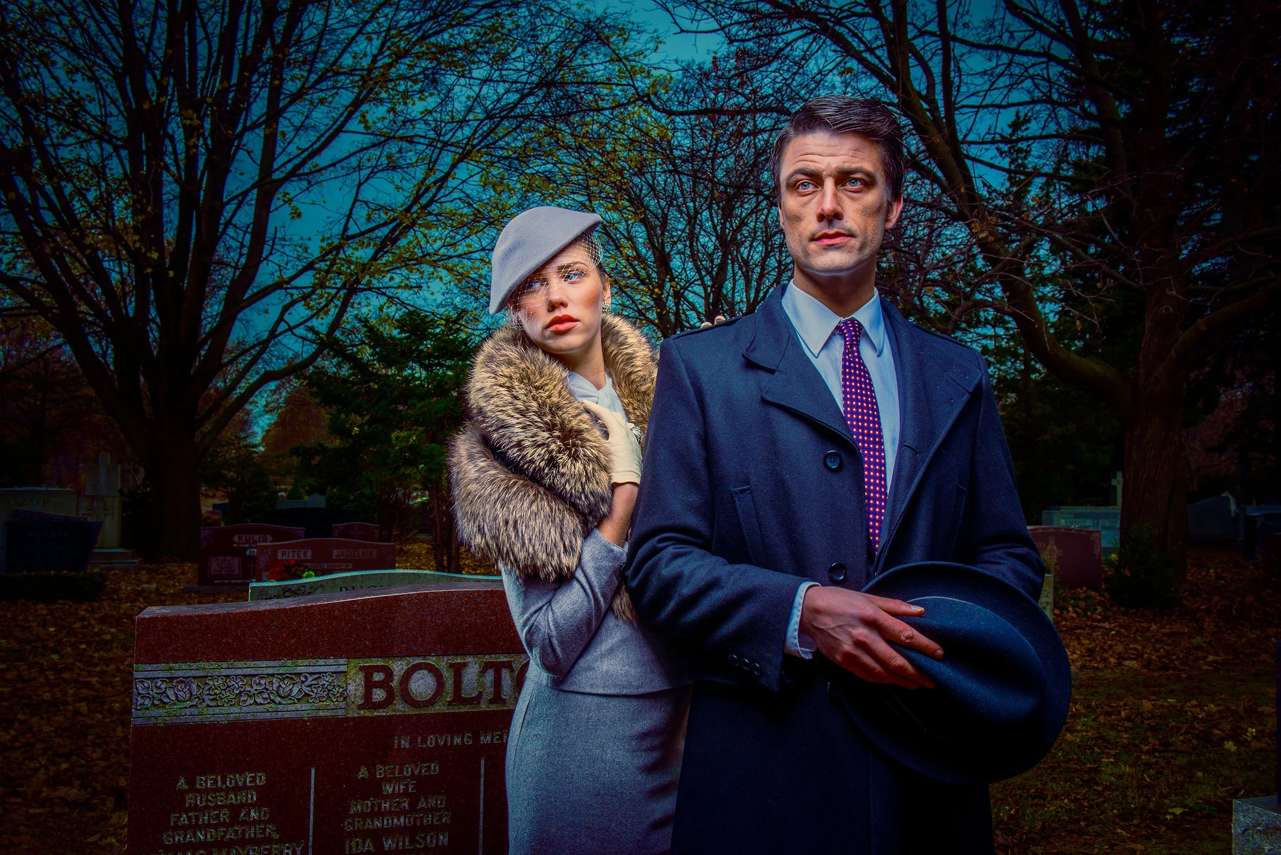 A man in a gray suit and a woman in a gray coat with a fur collar sit on a park bench in a cemetery. The woman wears a gray hat with a veil. They are surrounded by leafless trees and gravestones.