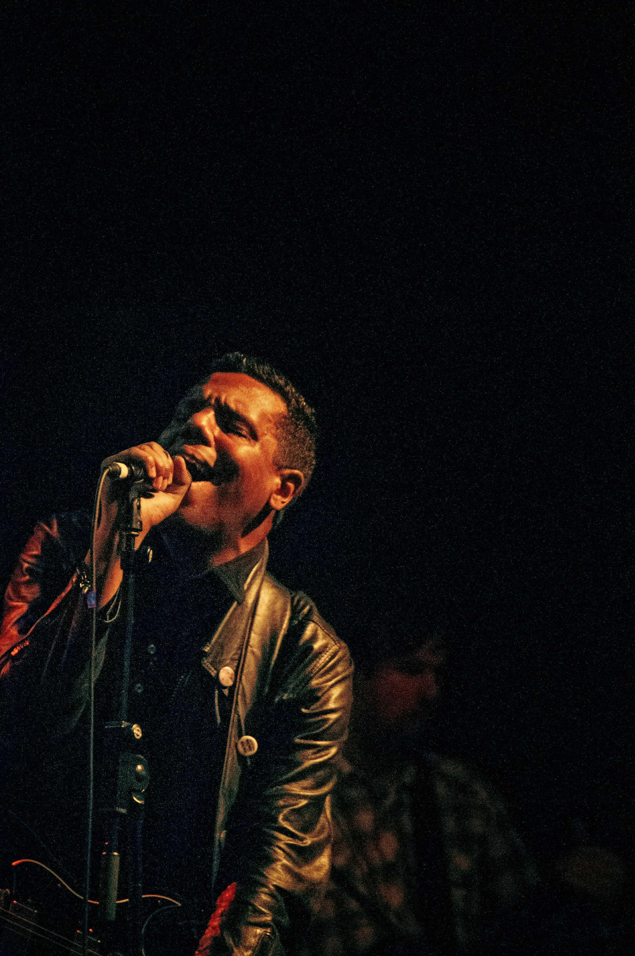 A young man singing into a microphone against a dark background with stars, wearing a leather jacket and holding the microphone with one hand.