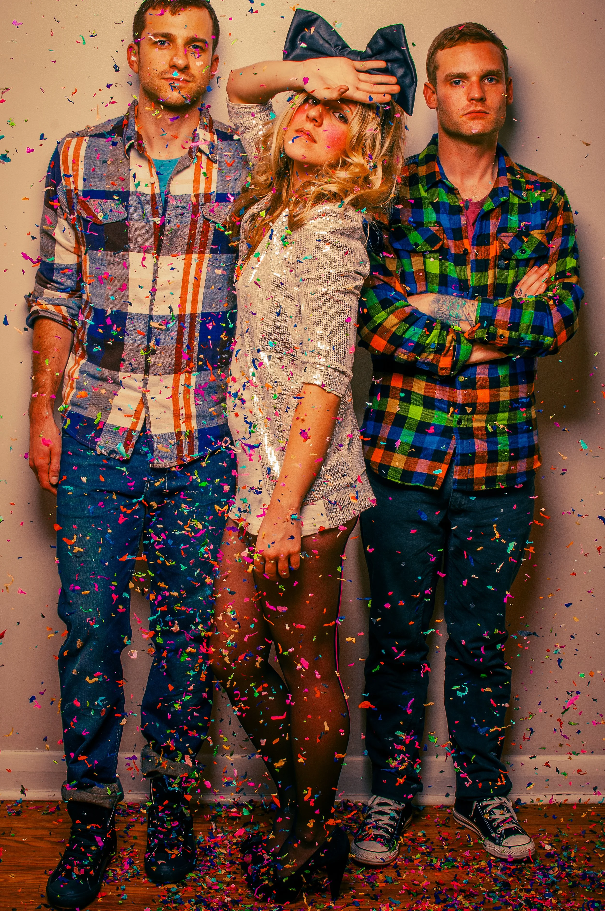 Three people standing against a wall with colorful confetti falling around them. The person in the middle is women with long blonde hair, wearing a large navy bow and a sparkly dress, striking a playful pose. The person on the left is a man with shor