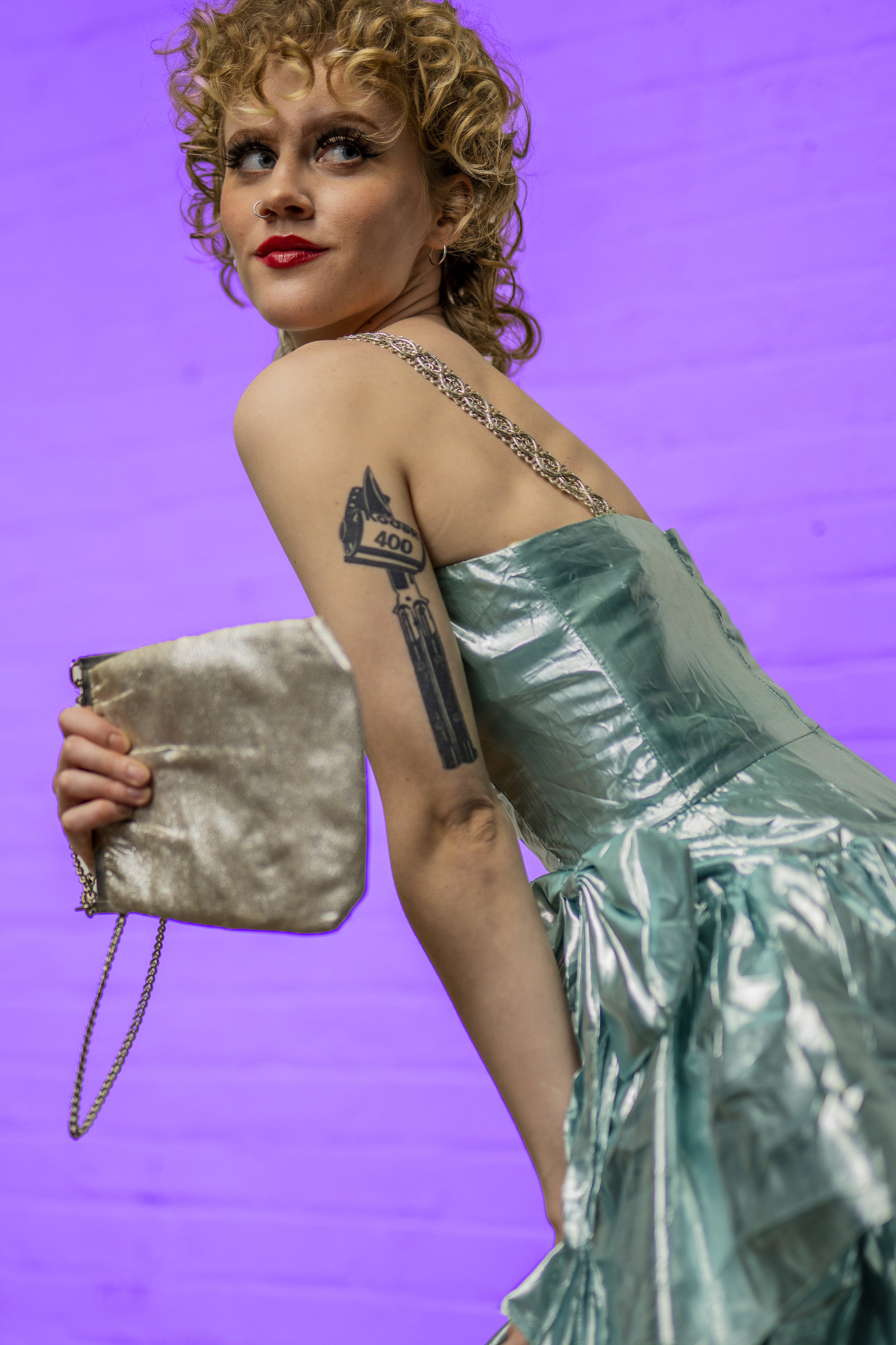 A woman with curly blonde hair, red lipstick, and a nose ring, dressed in a shiny, metallic dress, holding a gold velvet clutch with a chain, against a purple background.