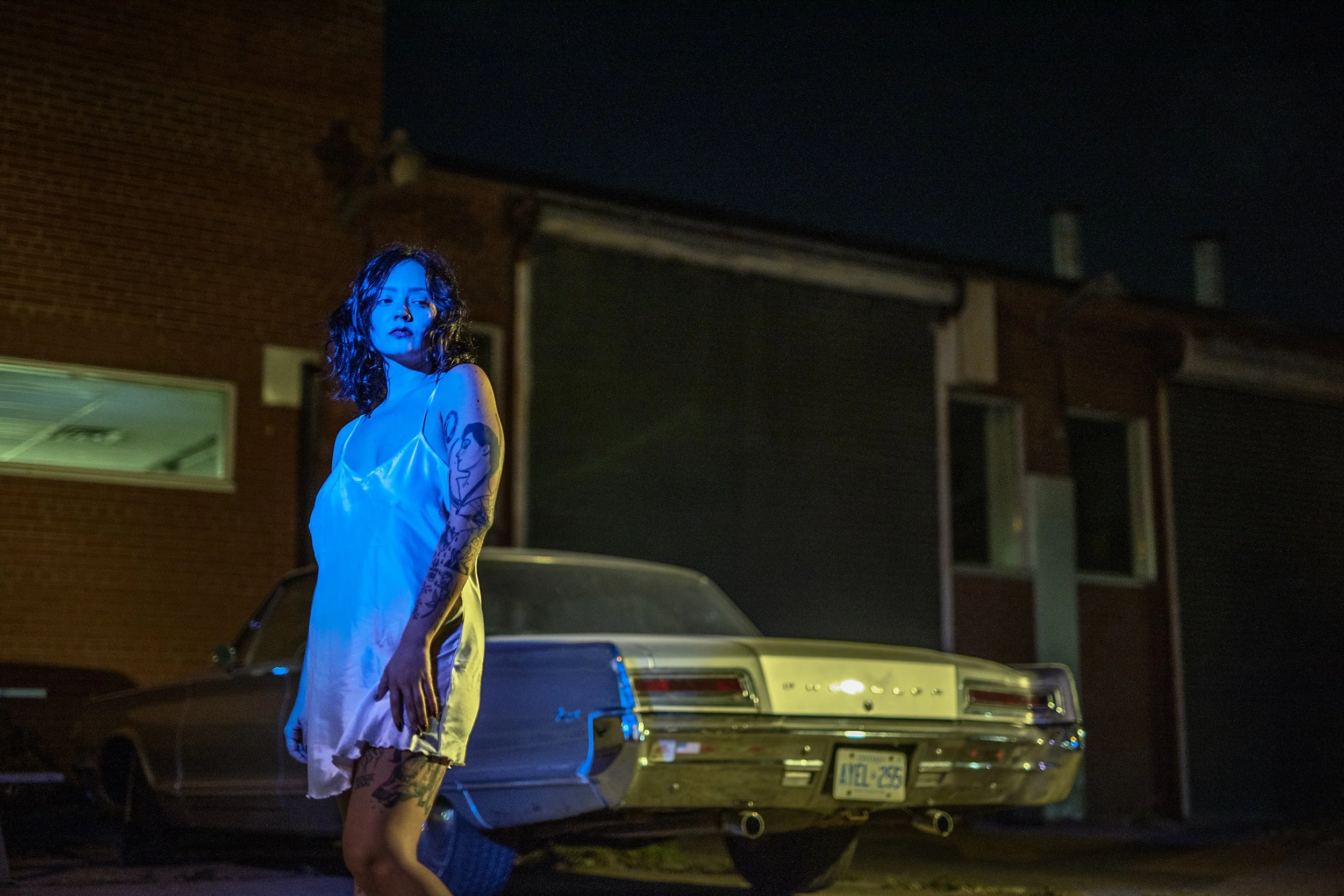 A woman with tattoos standing outside at night, illuminated by blue and yellow lights, with a vintage car and house in the background.