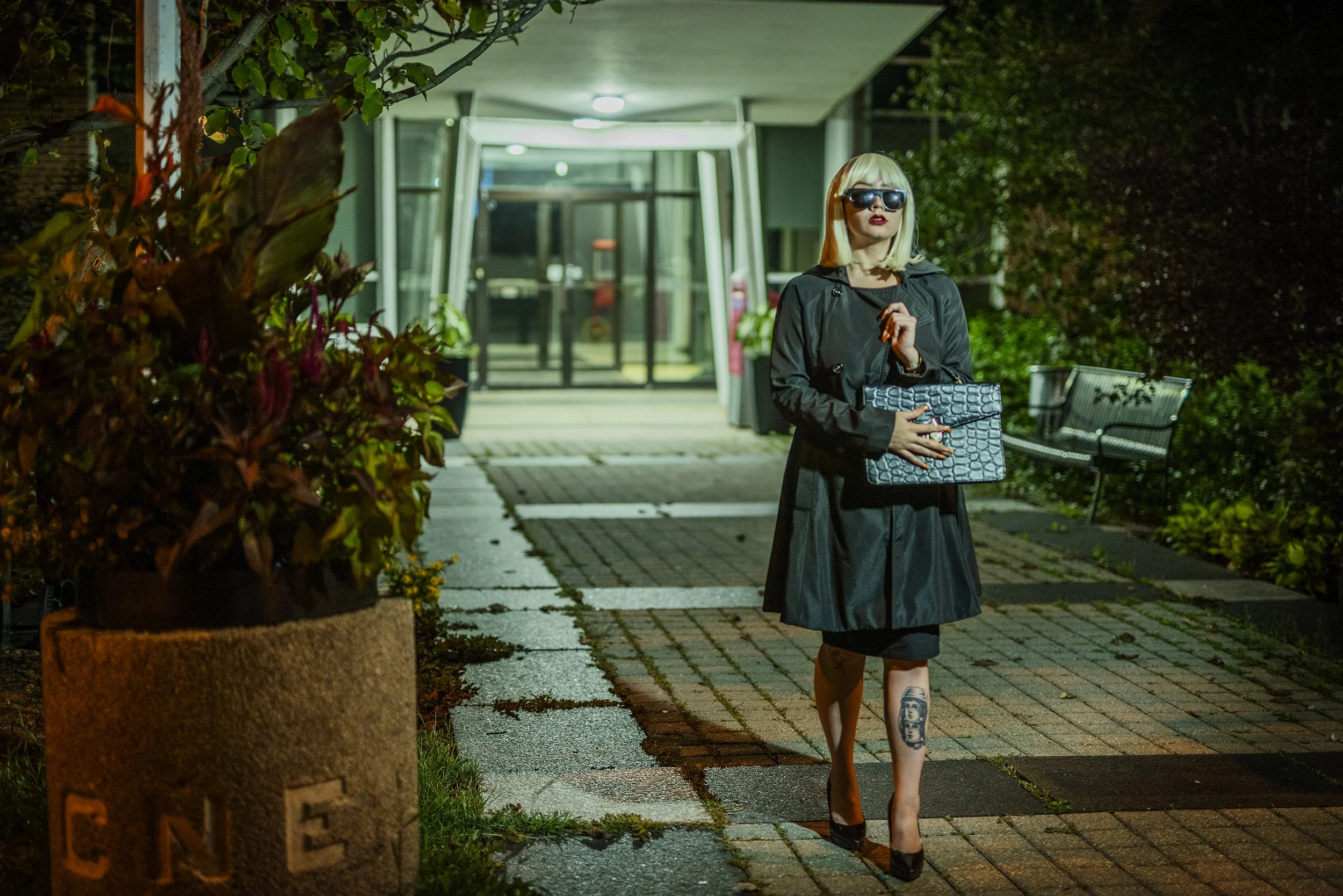 A woman with shoulder-length blonde hair, wearing dark sunglasses, a dark coat, and high heels, is walking on a dimly lit sidewalk at night while holding a patterned clutch. She has a tattoo on her left thigh visible as she walks.