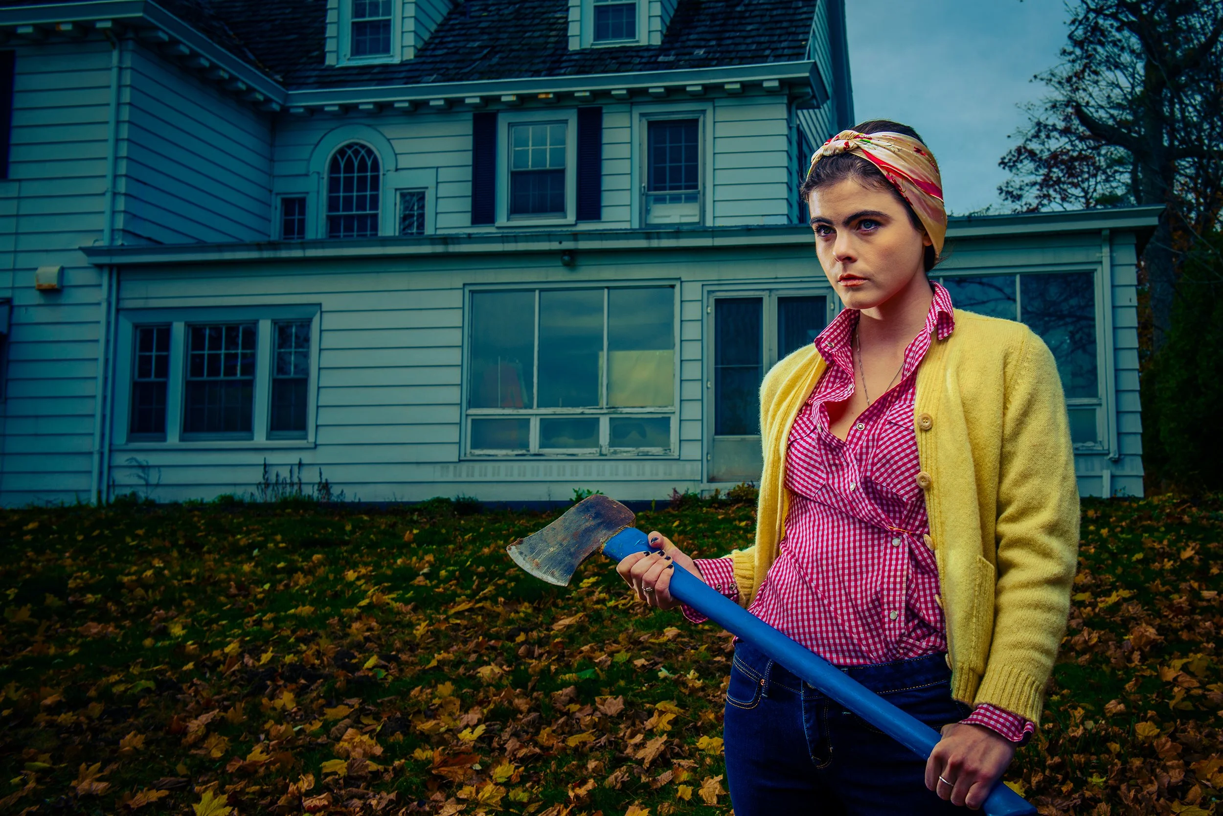 A woman with a colorful headscarf and yellow cardigan holding an axe stands in front of a large white house with multiple windows, surrounded by fallen autumn leaves.