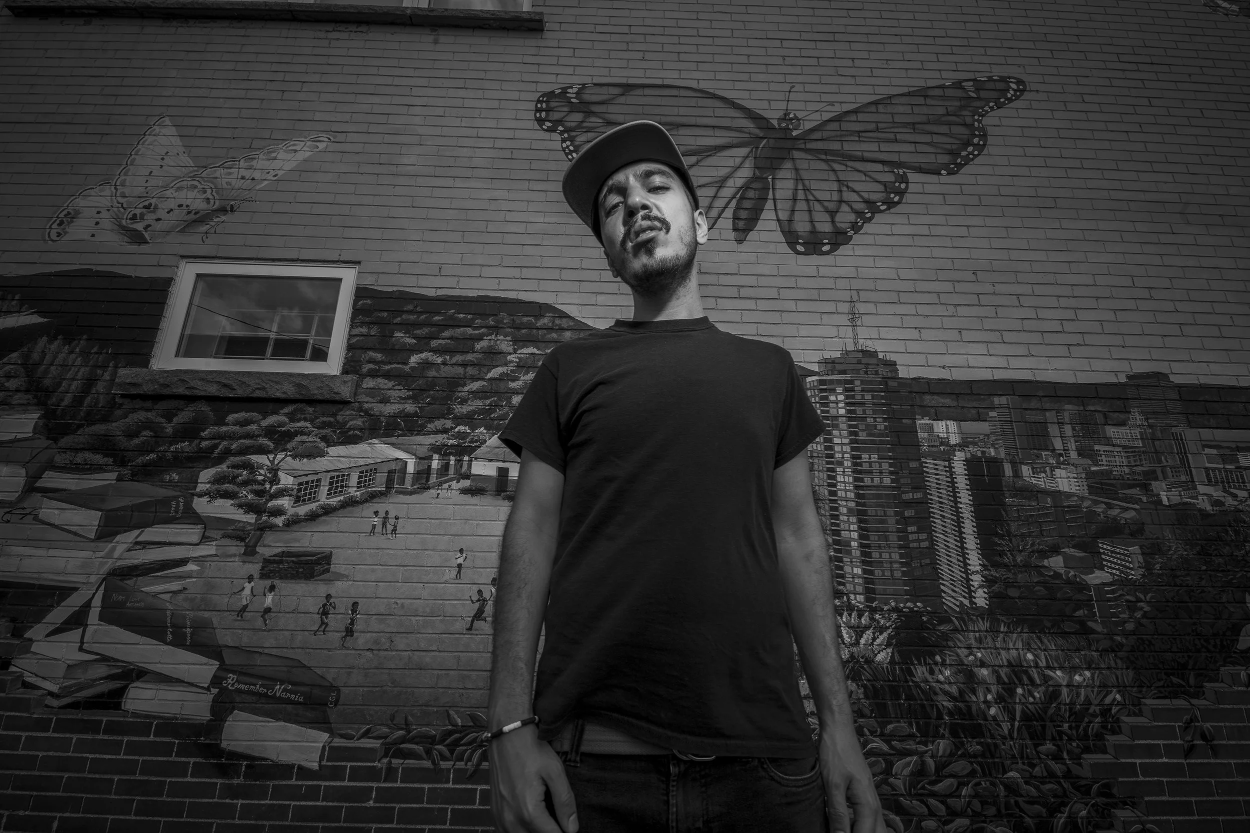 A black-and-white photo of a young man with a mustache and beard wearing a baseball cap and a black t-shirt, standing in front of a mural of butterflies and cityscape on a brick wall.