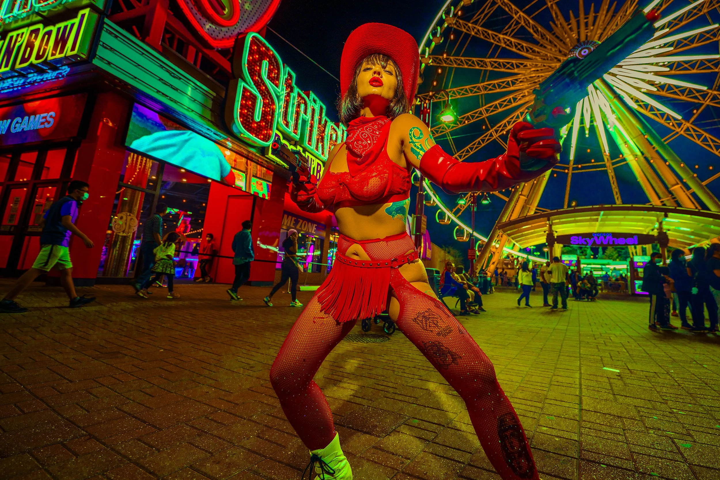 A person dressed in a red cowboy-themed outfit with tattoos, fishnet stockings, and a large red hat, posing with a toy gun at a brightly lit amusement park at night, with a colorful ferris wheel and neon signs in the background.