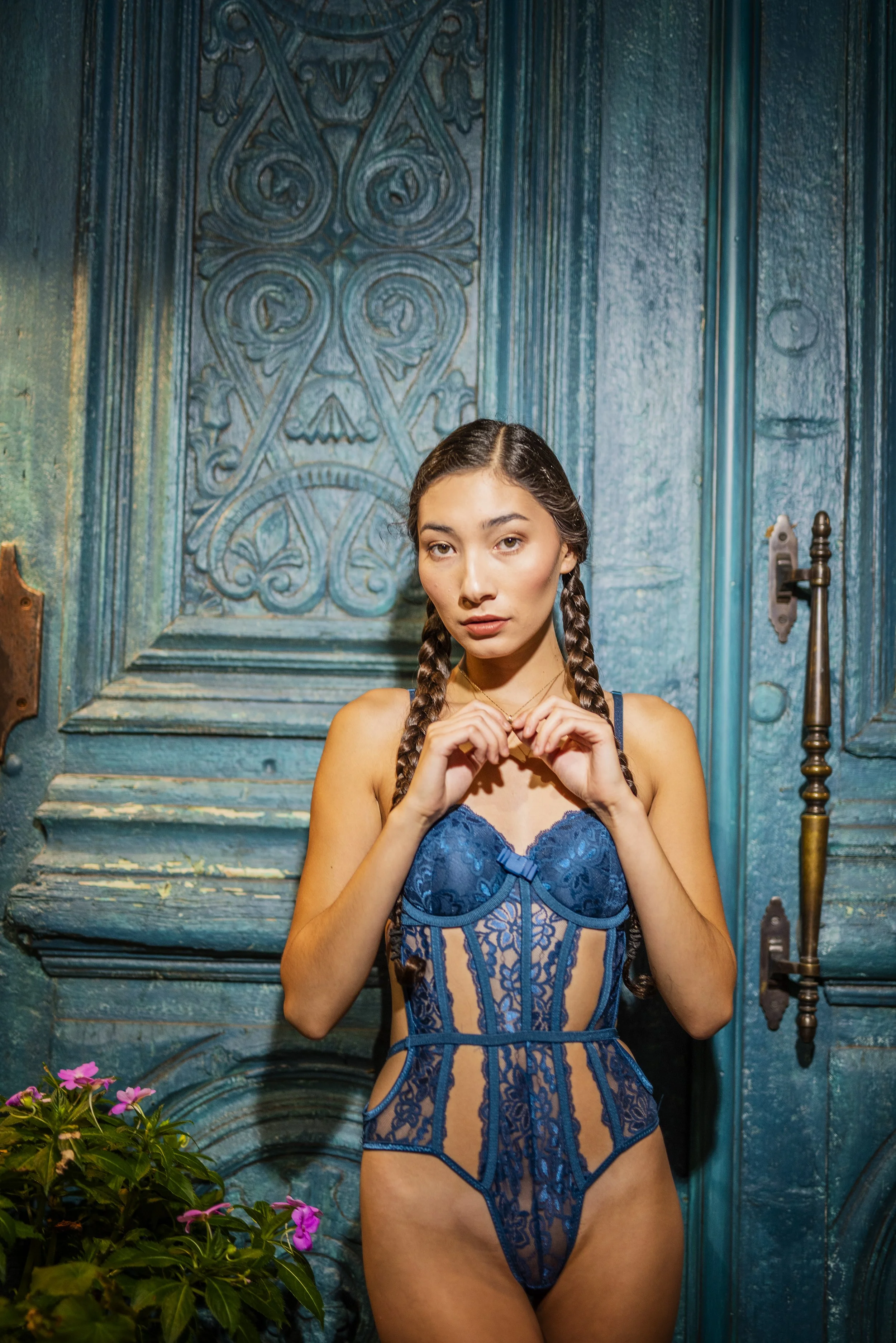A young woman with long dark hair in two braids wearing a blue lace lingerie bodysuit standing in front of a vintage blue door with intricate carvings and a handle. There are pink flowers and green foliage to her left.