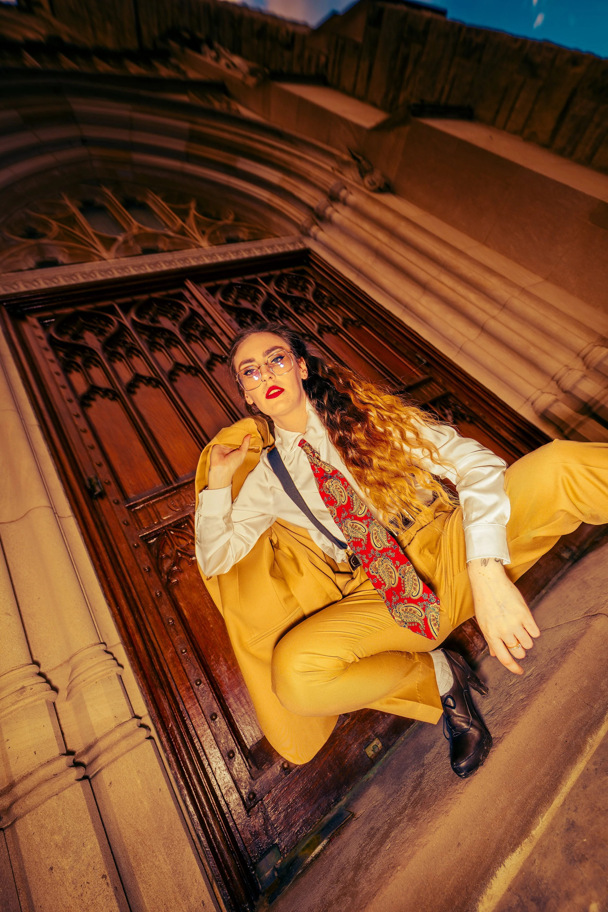 A woman with long curly hair, wearing glasses, a striped tie, and a beige blazer, sitting on the ground with her leg bent, outside a historic building with arched wooden doors and stone architecture, during sunset or twilight.