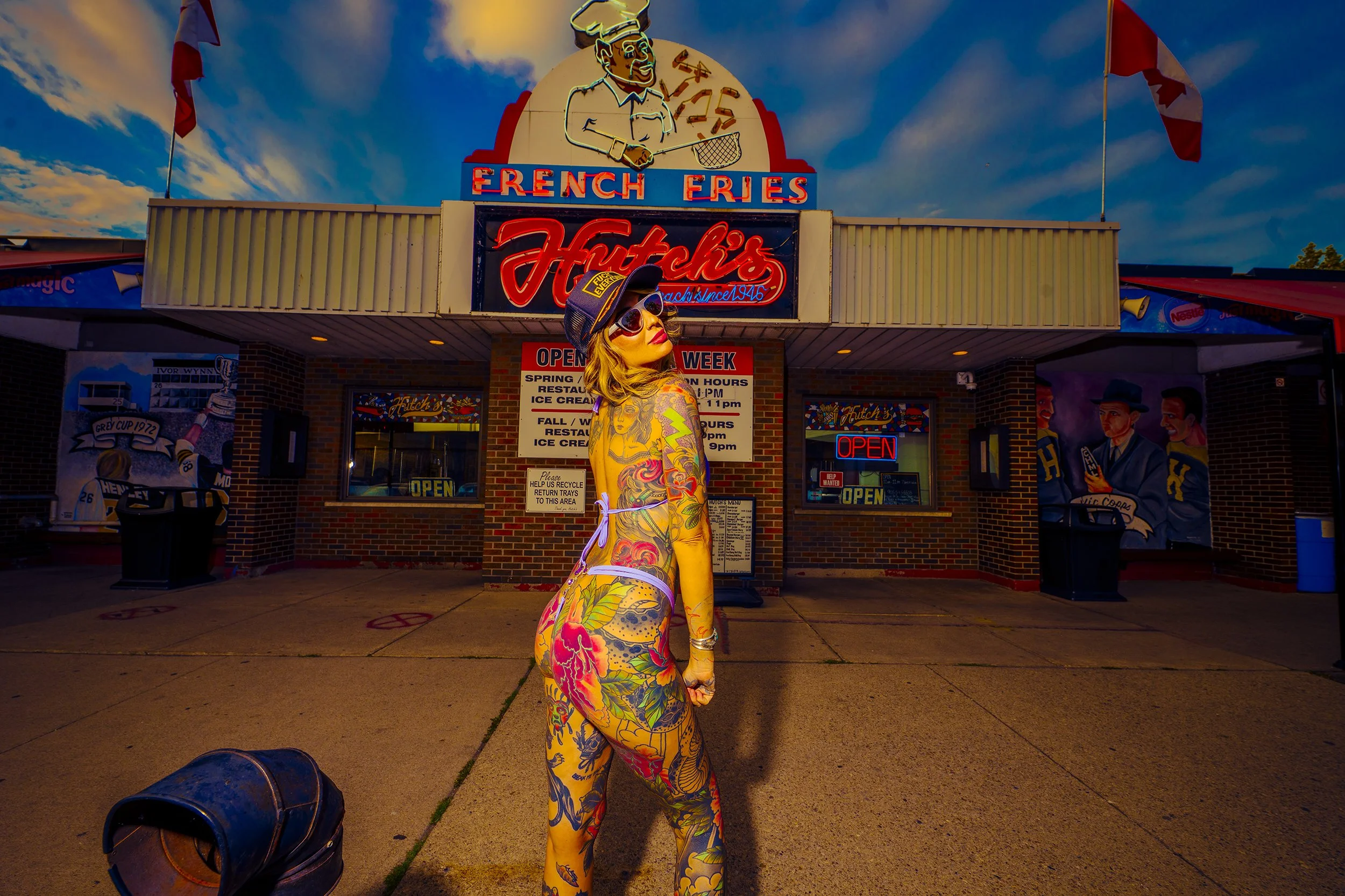 A woman with colorful tattoos wearing sunglasses, a cap, and a swimsuit, standing in front of Fitch's ice cream shop at sunset.