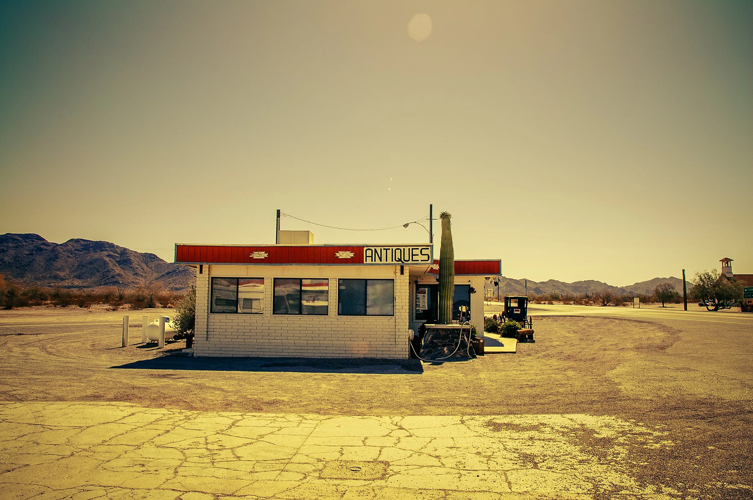 A small white brick building with a red sign that reads 'ANTIQUES' and a cactus outside, situated in a desert landscape with mountains in the background and a clear sky.