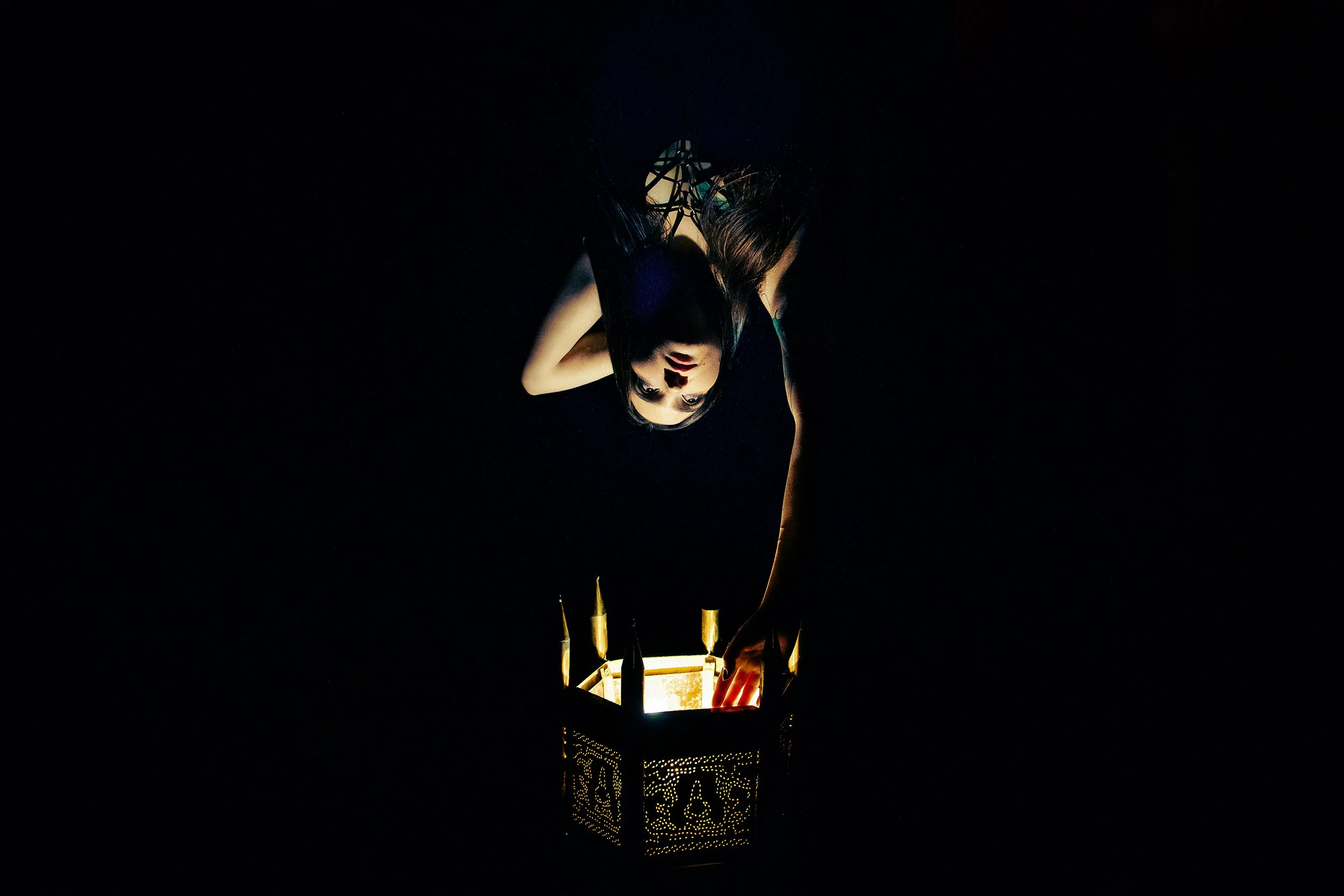 A woman in a black dress is leaning over a lantern-like structure with intricate patterns, lit from within, illuminating her face. The scene is dark with the focus on the glowing lantern.
