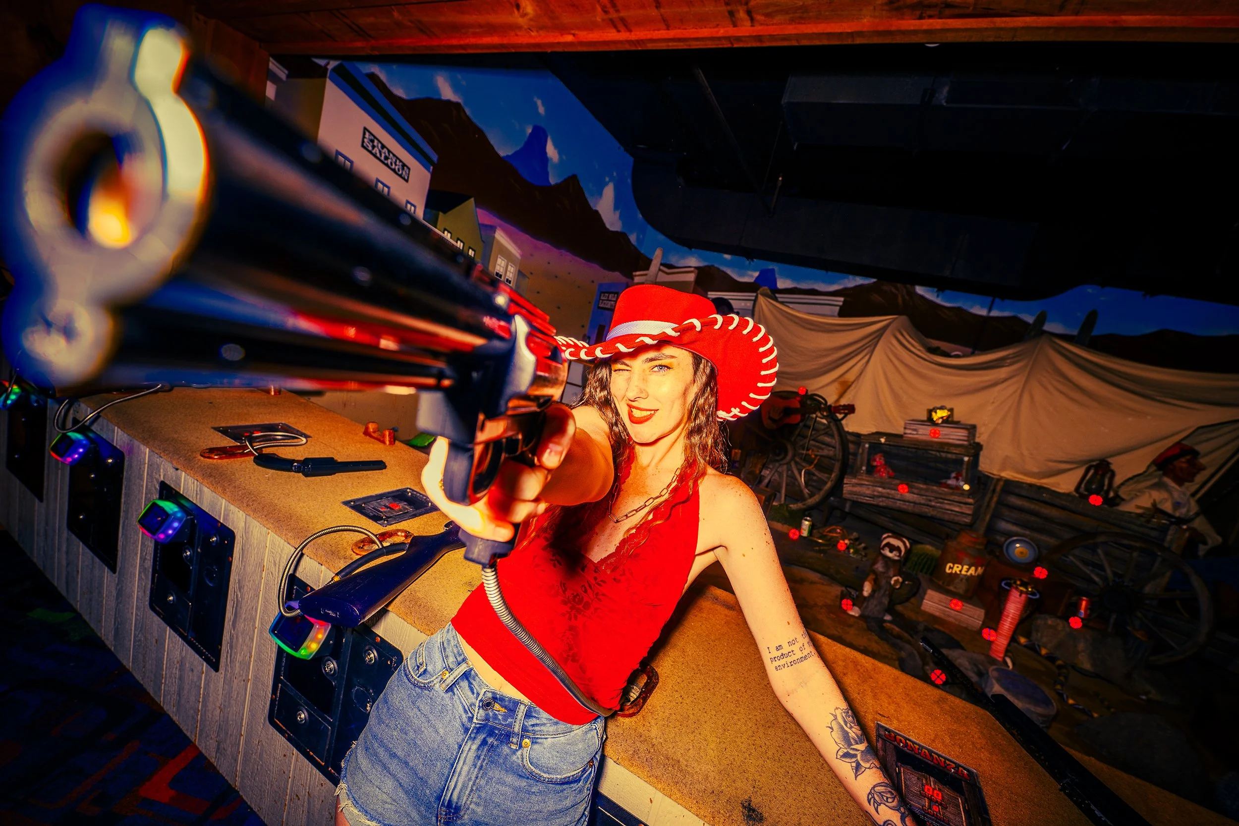Woman wearing a red cowboy hat and red top pointing a gun towards the camera inside a colorful room with mountain mural and various objects on the counter.