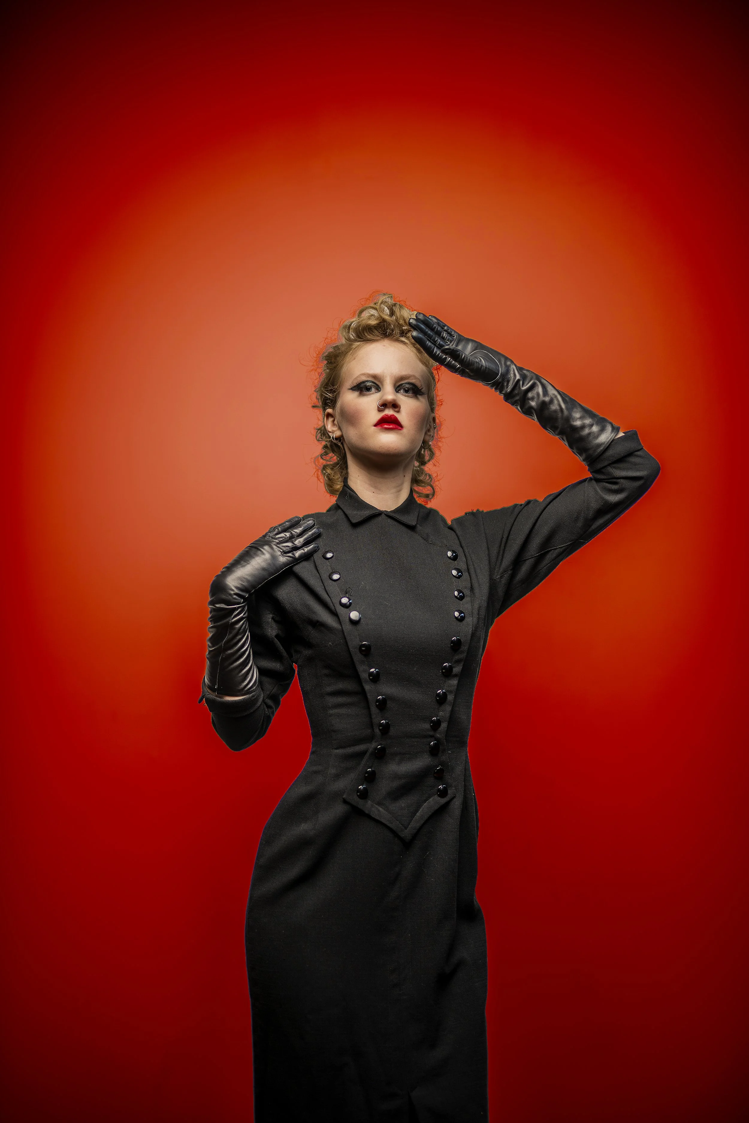 A woman dressed in a vintage black dress and long black gloves, with blond curly hair, posing against a red background.