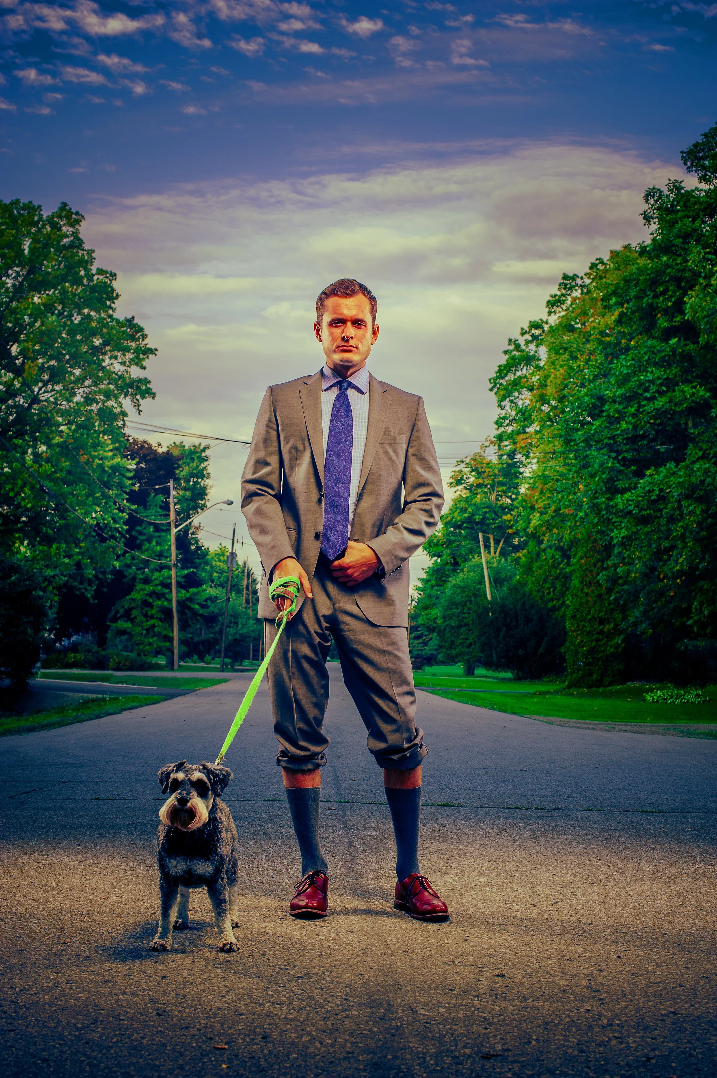 A man in a gray suit, red shoes, and a tie stands on a street holding a leash attached to a small dog. The man looks serious, and the dog appears to be a small breed with black, white, and gray fur. The background shows trees, utility poles, and a cl