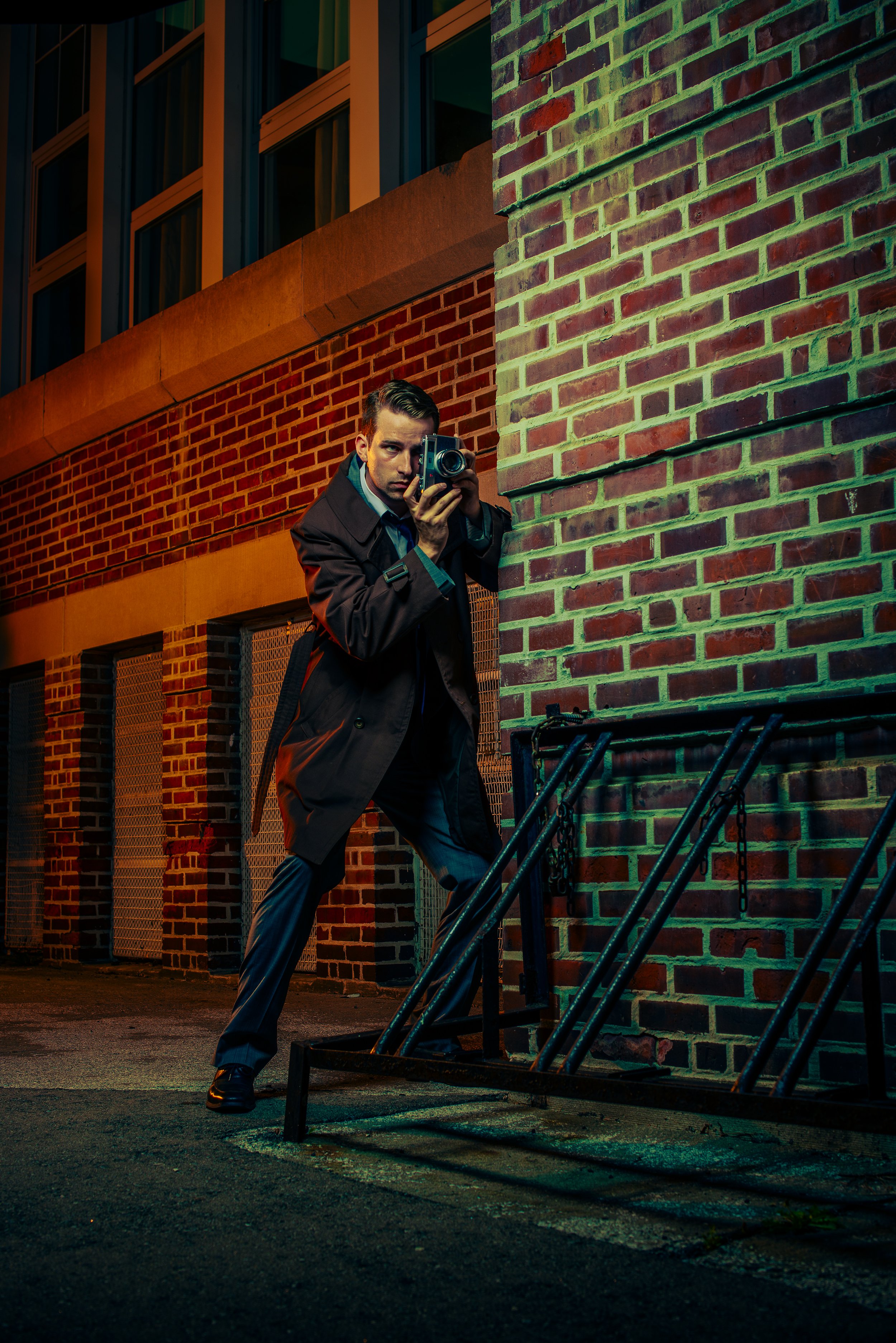 A man in a black coat and dress pants holding a camera, standing behind a metal barrier in an urban, brick-walled environment at night.
