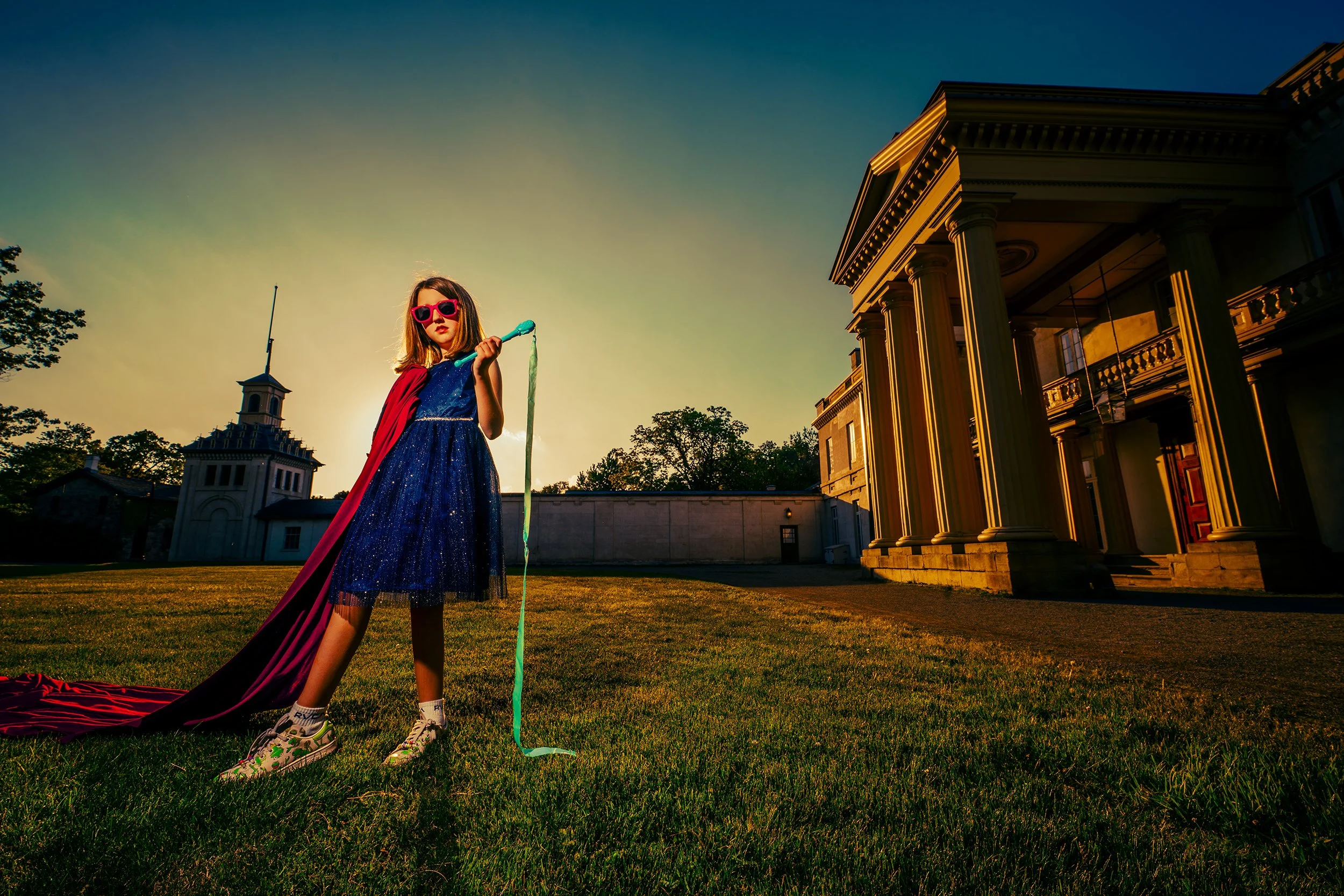 A young girl dressed as a superhero with a cape and sunglasses stands on a grassy field at sunset holding a streamer. Behind her are a large classical building with columns and a smaller building with a spire on the roof, with trees and a clear sky i