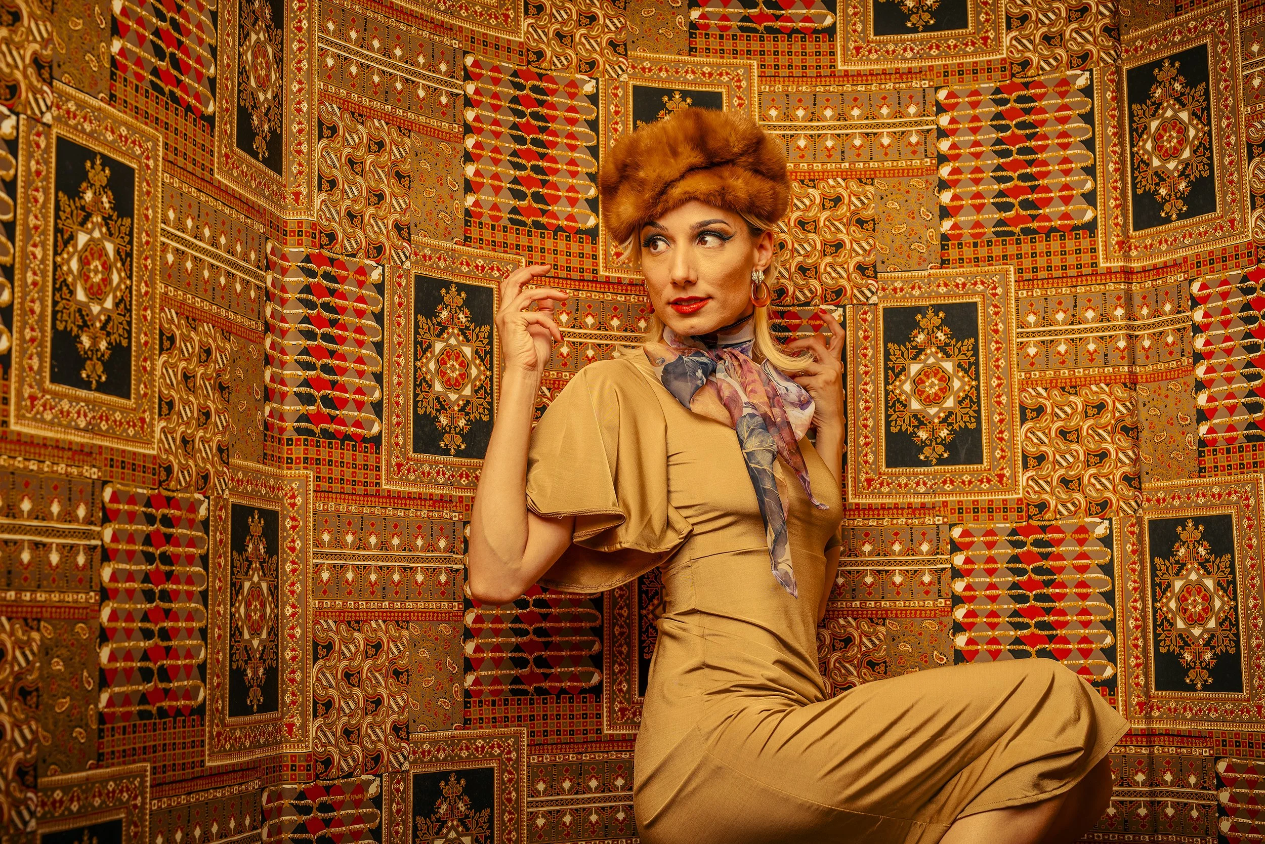 A woman wearing a brown dress, a colorful scarf, and a large fur hat, sitting against a backdrop of ornate, patterned gold, red, black, and cream wallpaper.