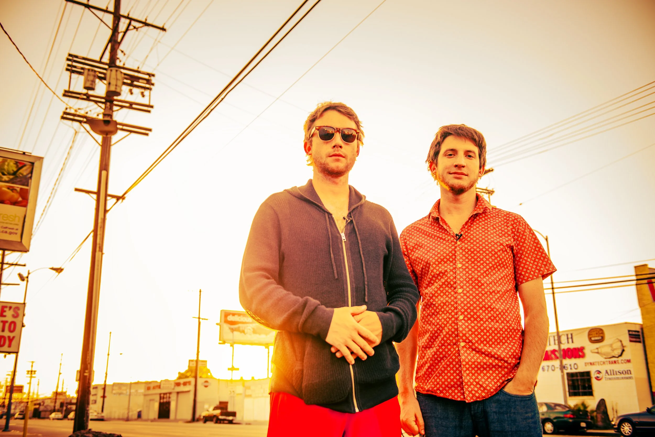 Two young men standing outdoors during sunset, with power lines and signs in the background. One wears sunglasses and a hoodie, the other wears a patterned short-sleeve shirt.