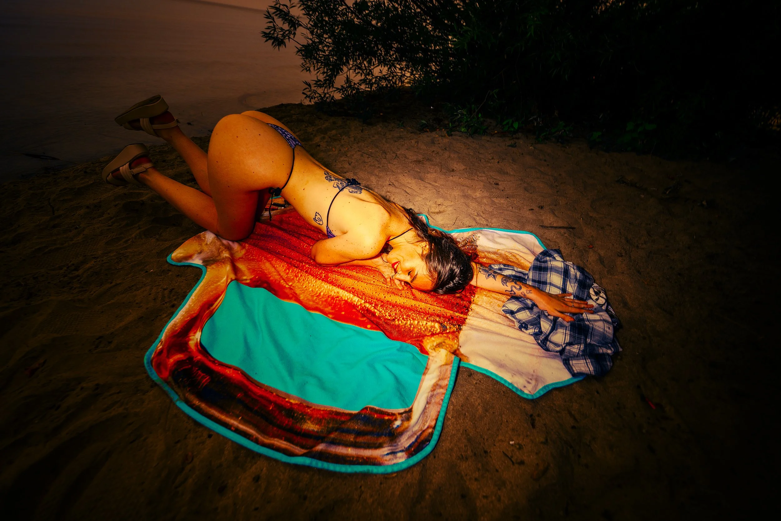 A woman with tattoos and long hair lying on her stomach on a beach towel, wearing a bikini, with her head resting on her arm, at night with plants and darkness surrounding her.