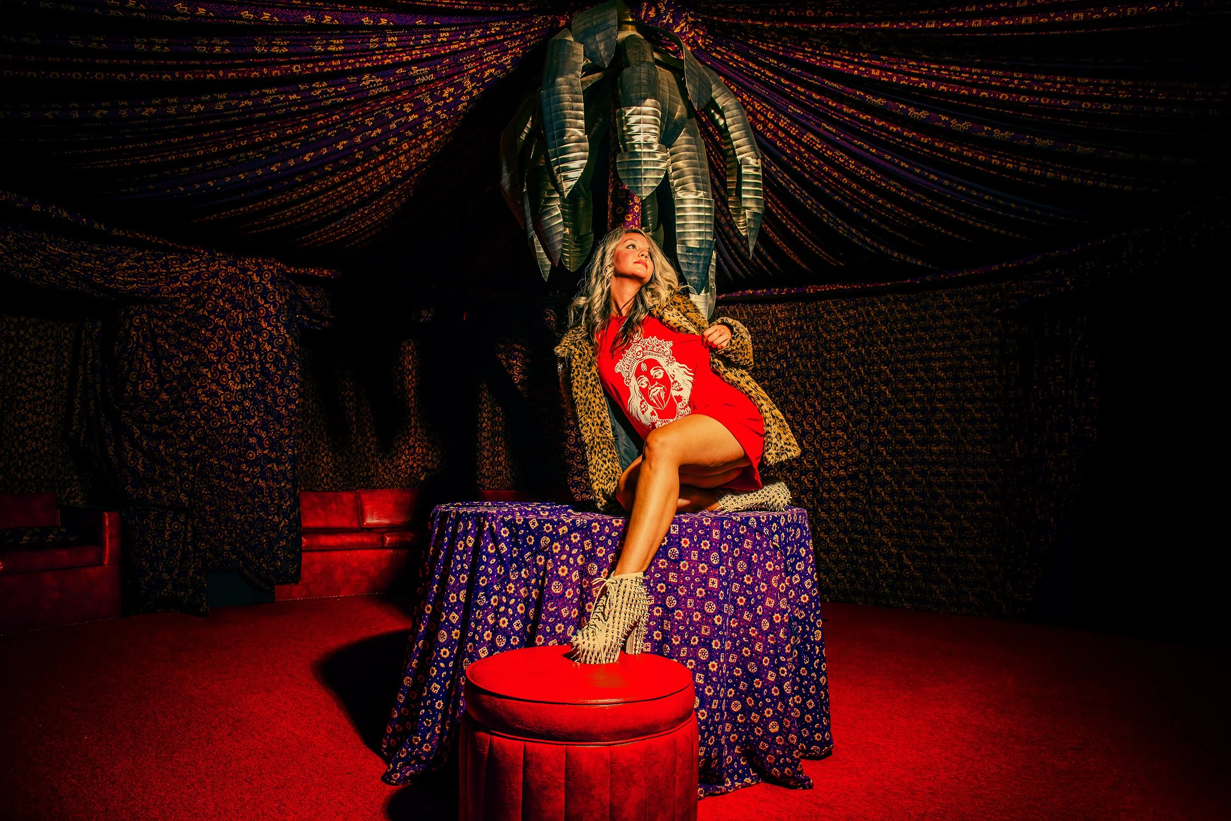 A woman with wavy blonde hair sitting on a table in a colorful, decorated room with blue patterned fabric covering the table and walls, and draped fabric on the ceiling. She is wearing a red dress with a graphic lion design, a leopard print coat, and