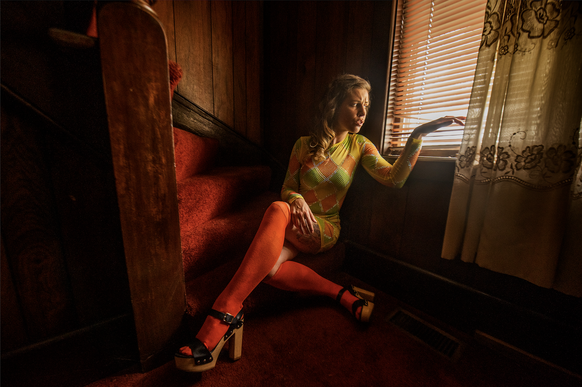 A woman sitting on a red-soaked vintage staircase by a window with blinds and lace curtains, wearing a colorful geometric patterned dress, orange thigh-high stockings, and black platform heels.