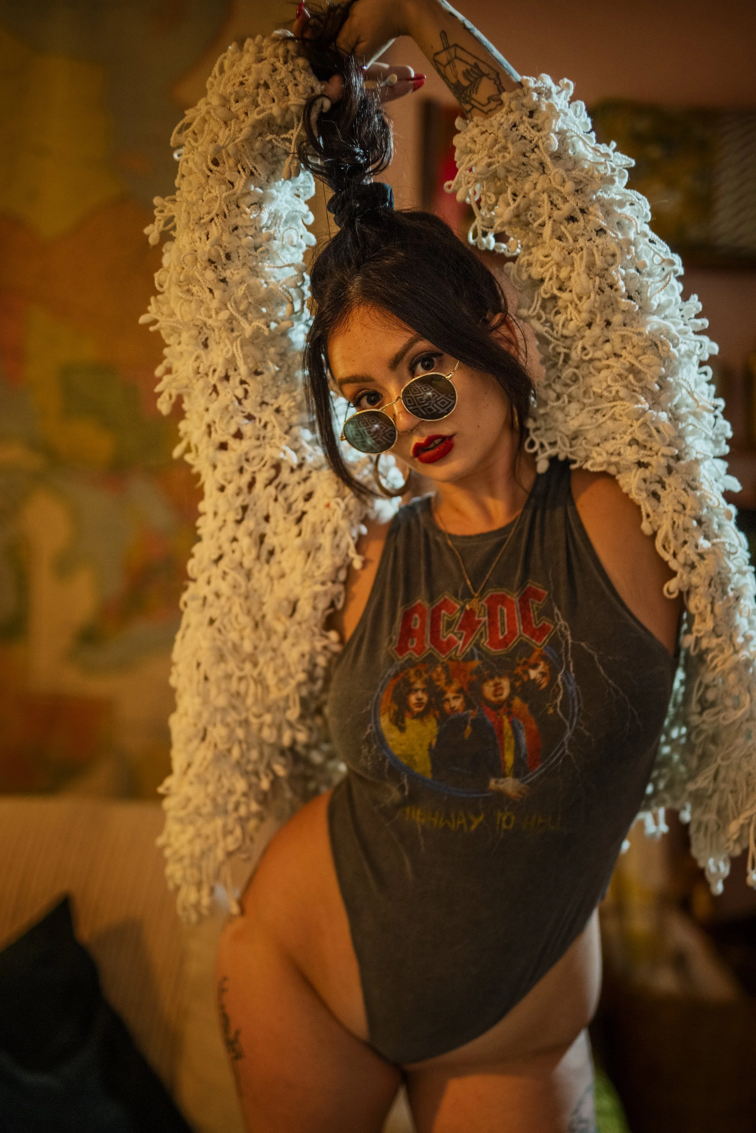 A woman wearing sunglasses and a sleeveless AC/DC band t-shirt poses indoors, holding her hair up with one hand and looking at the camera. She has red lipstick, dark hair, and tattoos on her arms and thigh, with a crocheted cardigan draped over her s