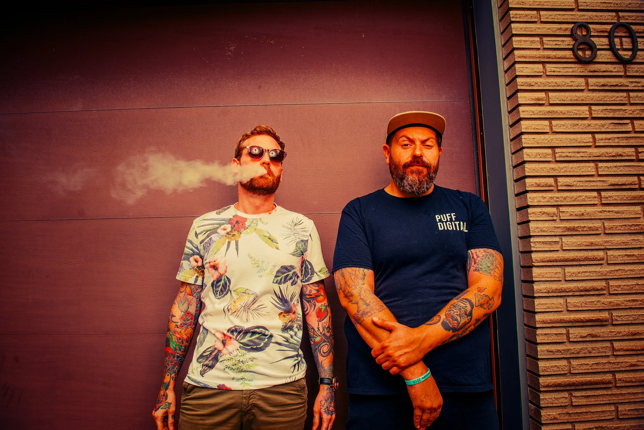 Two men with tattoos standing against a red wall next to a brick corner, one exhaling vapor and wearing sunglasses and a floral shirt, the other with a beard, a cap, and a black t-shirt that says 'PUFF DIGITAL'.