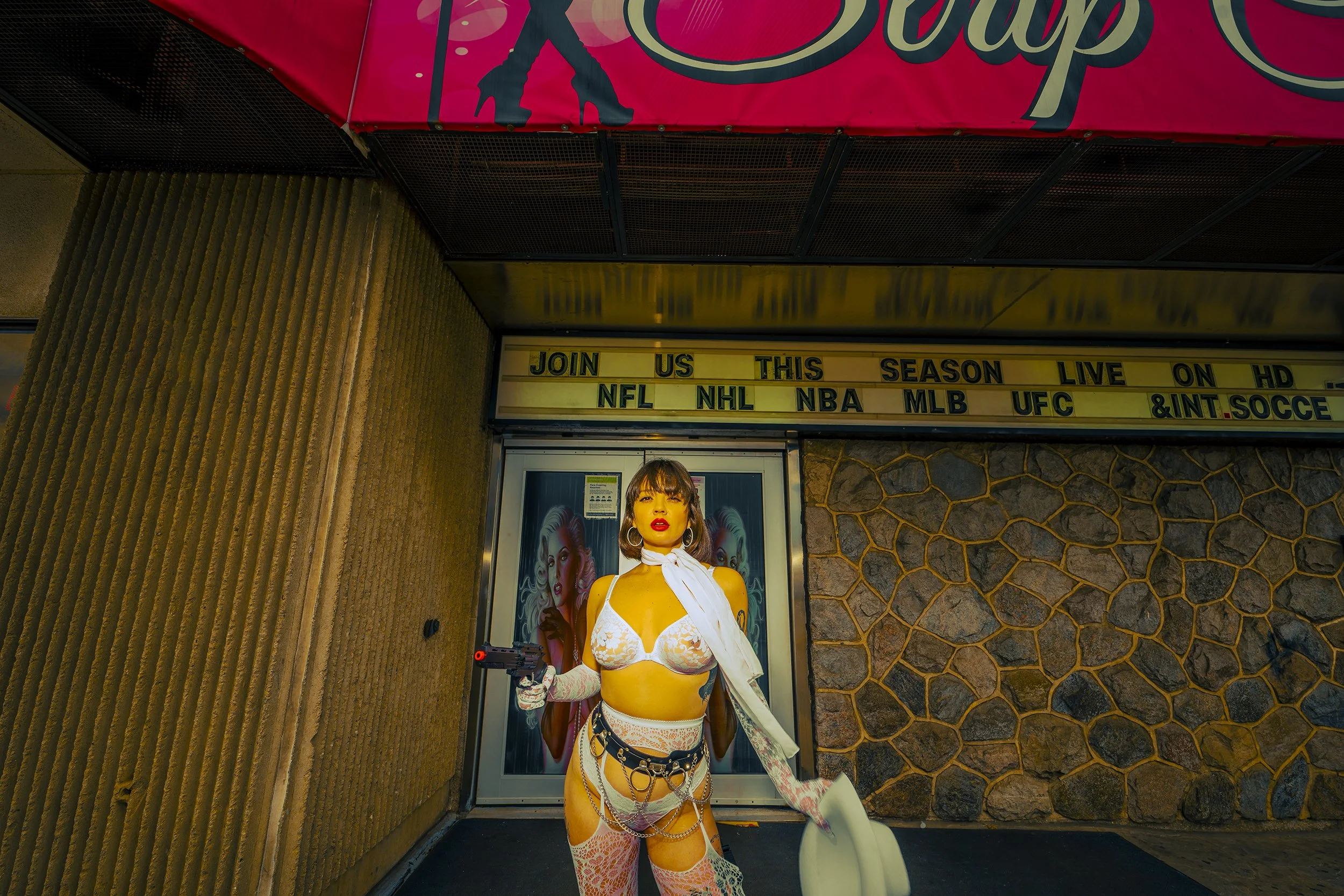 A woman stands outdoors in front of a movie theater entrance, holding a gun, dressed in a provocative outfit with chains and lace, with a poster of a woman behind her.