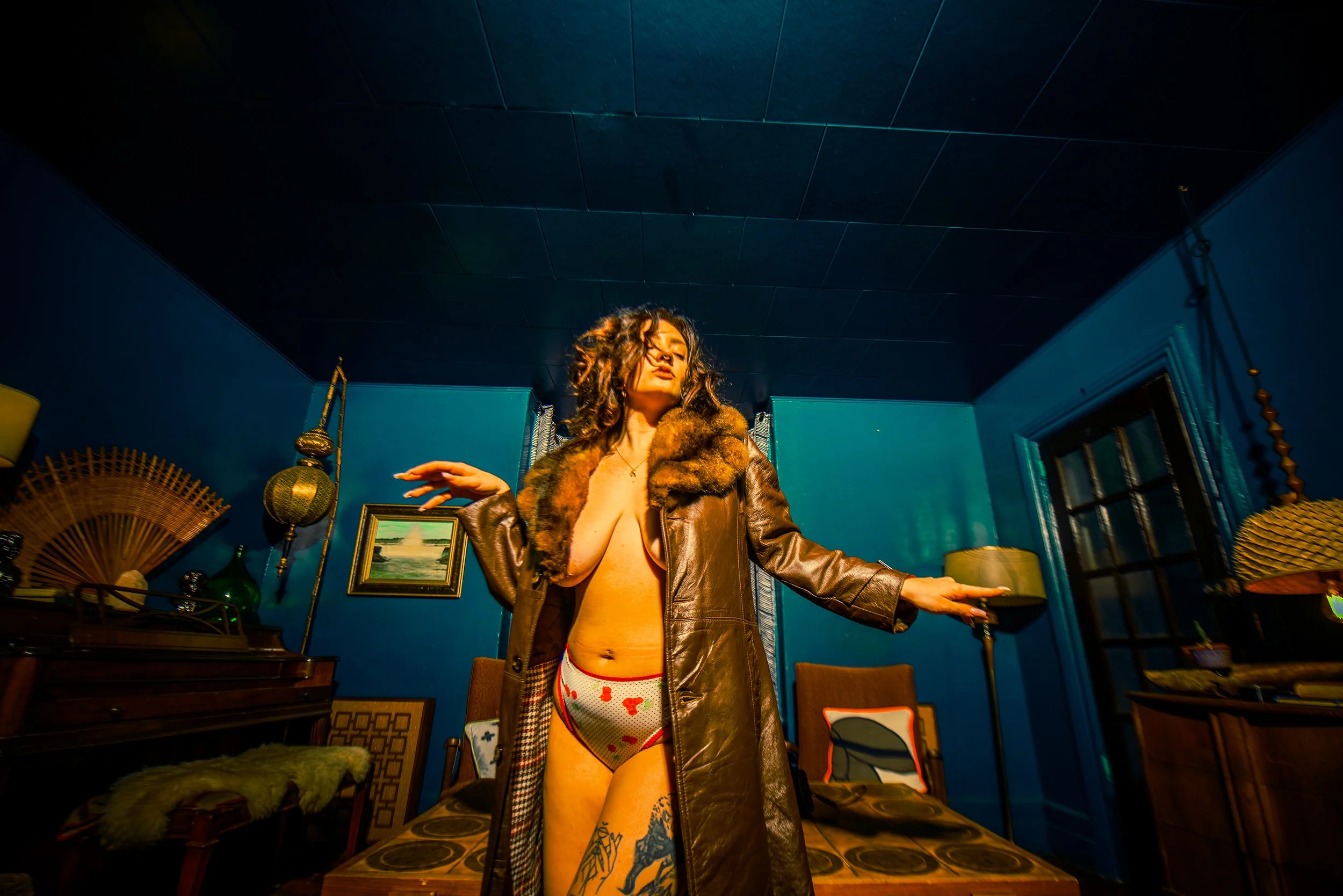 A woman with long curly hair standing in a teal-colored room, wearing a brown leather jacket with a fur collar, and patterned underwear. She has one arm extended and appears to be posing.