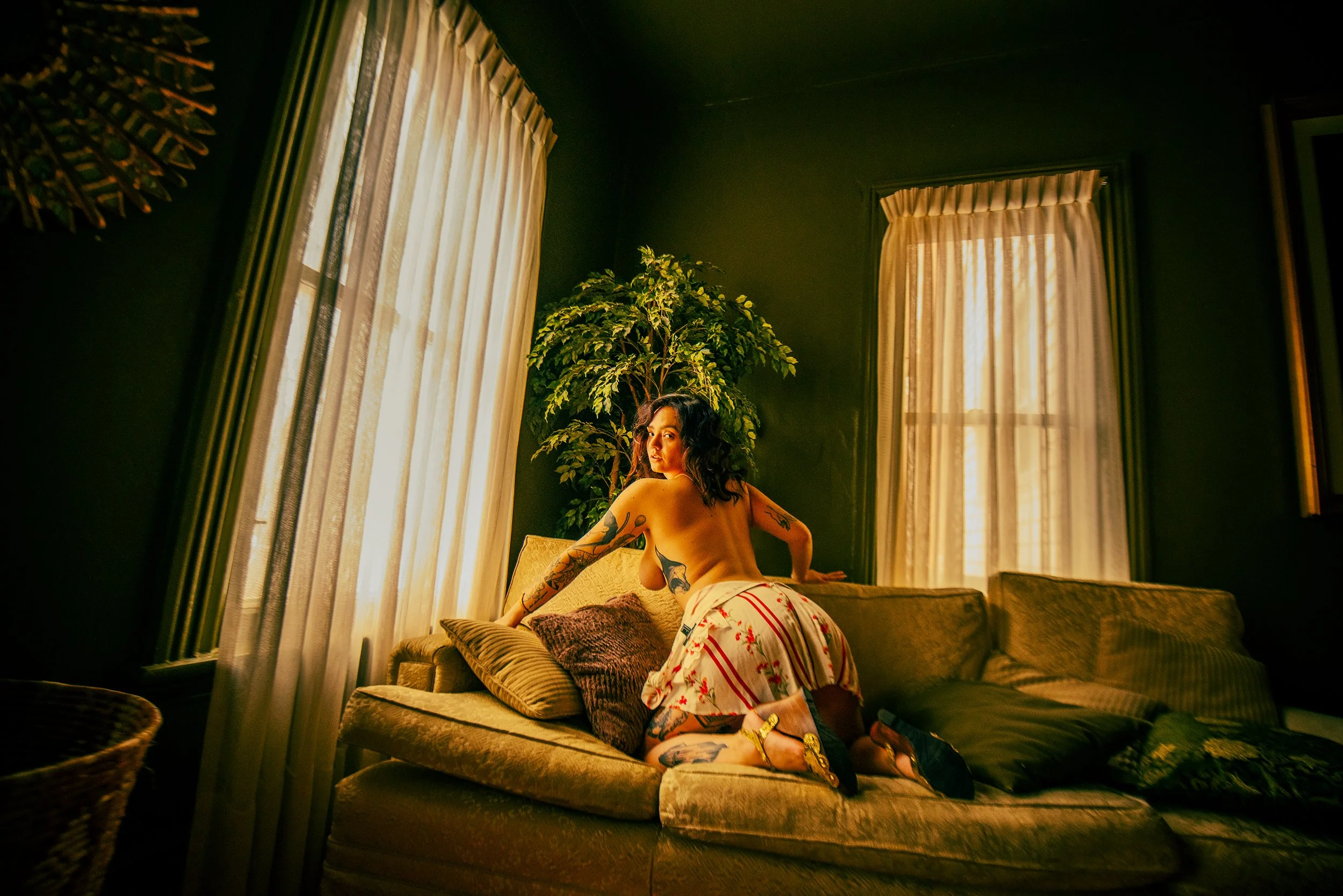 A woman with tattoos relaxing on a couch in a dimly lit room with large windows covered by curtains and a large potted plant.
