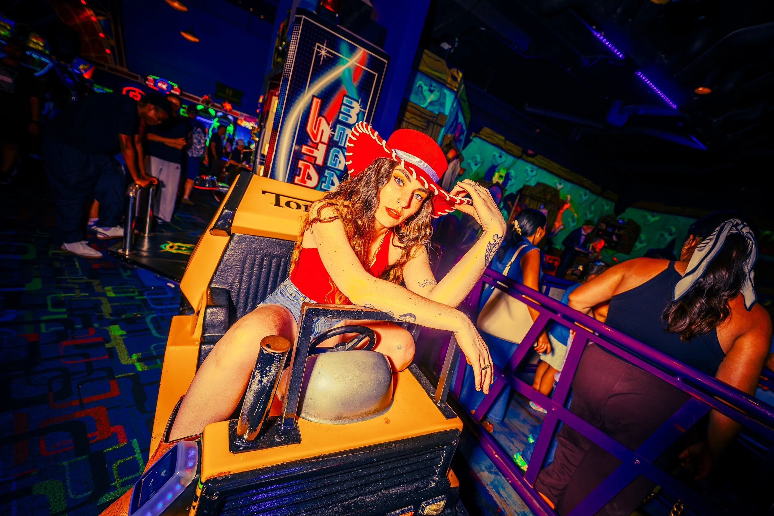 A young woman with wavy hair and tattoos, wearing a red top and a red sombrero, sitting in a yellow bumper car in an arcade with neon lights and other people playing games in the background.