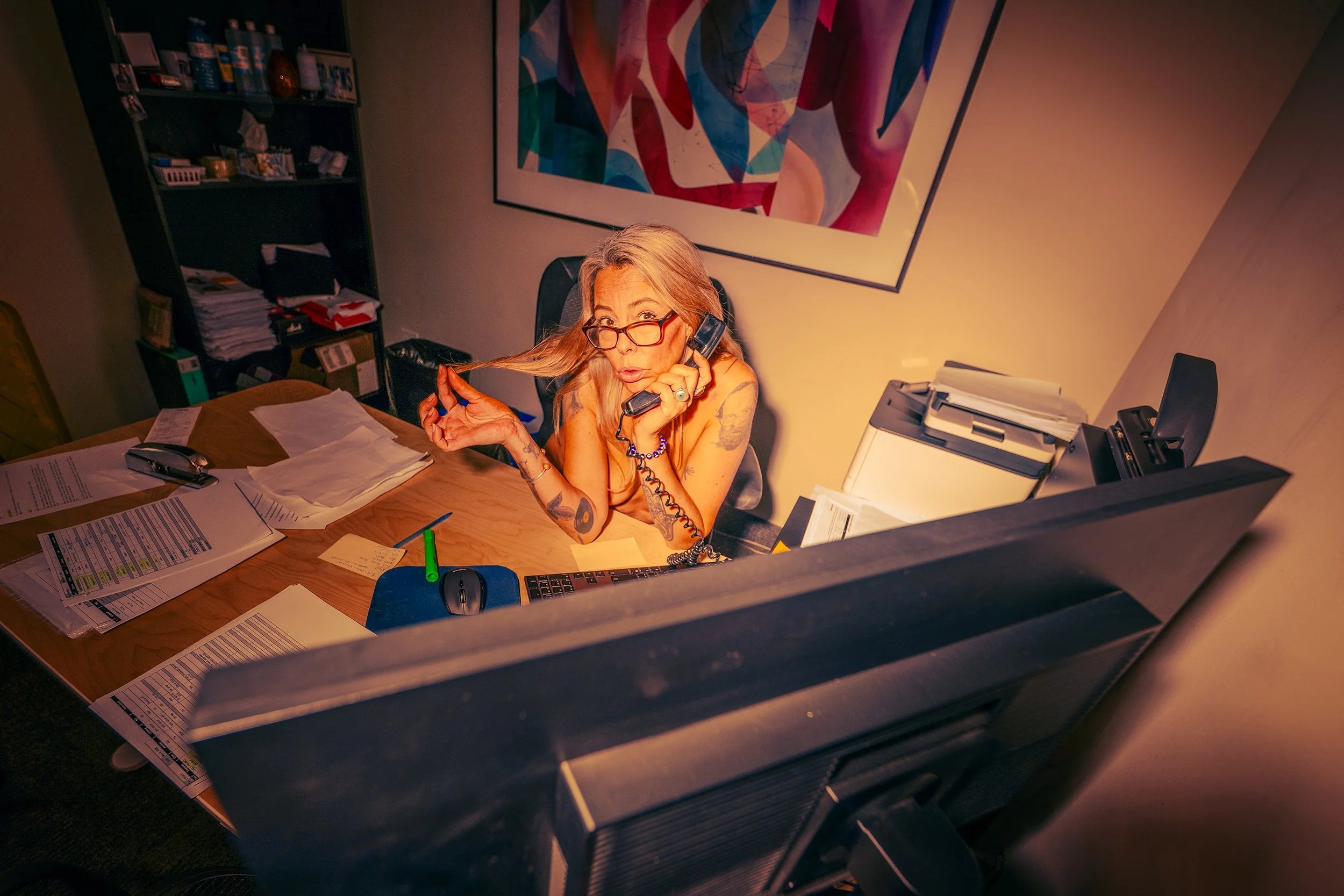 A woman with glasses and long hair sitting at a cluttered desk, talking on the phone, with papers, a keyboard, and office supplies. There is a colorful abstract painting on the wall behind her.