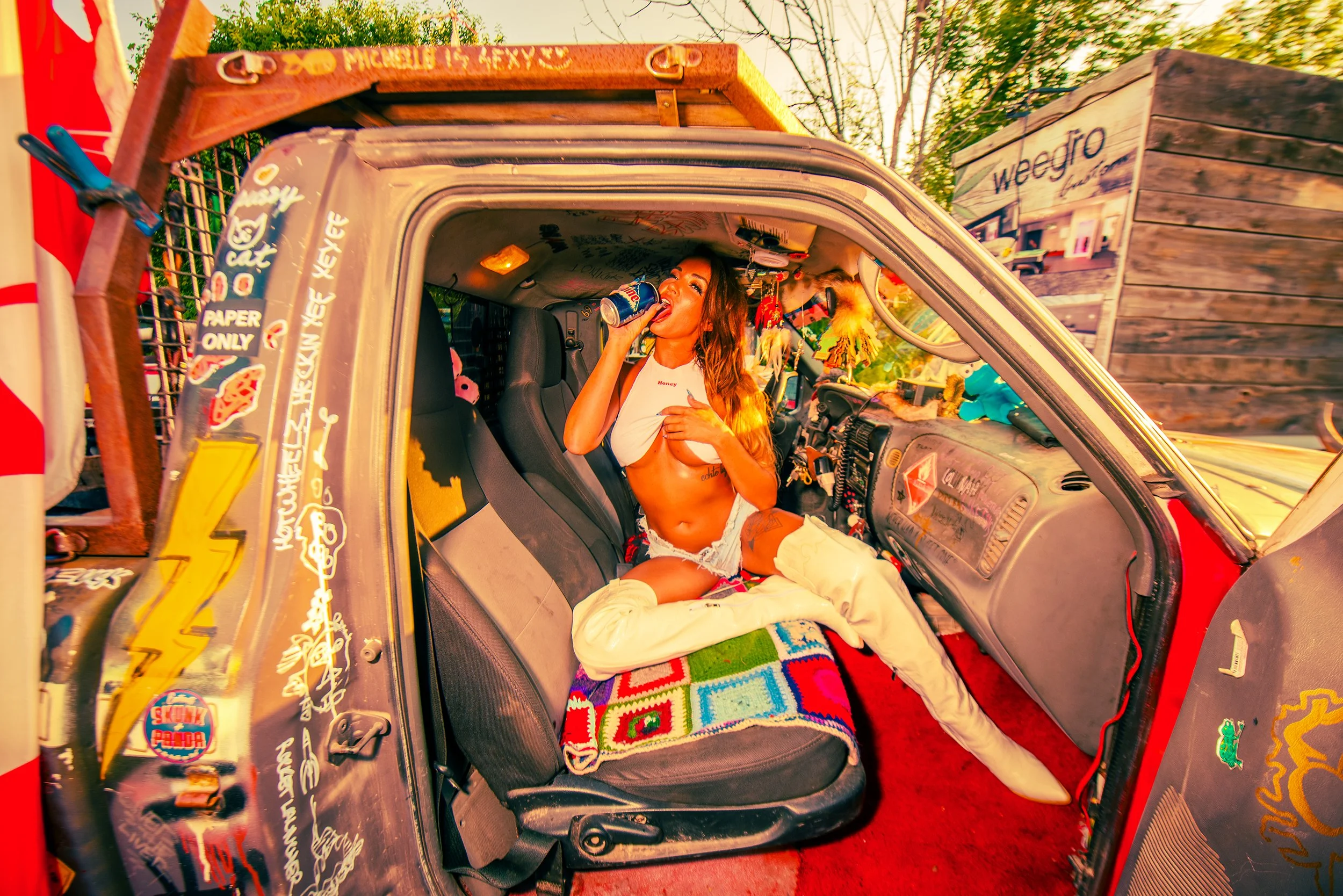 A young woman with long hair, wearing a white crop top and shorts, is sitting in the passenger seat of a decorated truck or van, drinking from a soda can and smiling. The vehicle has various stickers, decals, and colorful blankets inside, with a rust