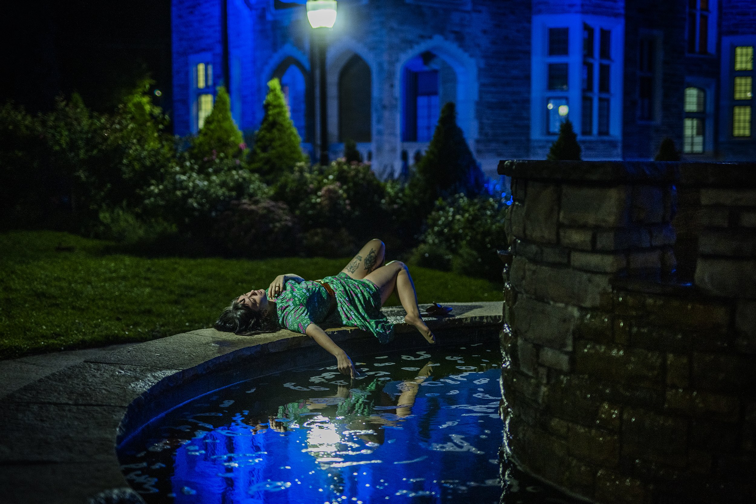 A woman laying on a small bridge at night, smiling, wearing a colorful dress, with a fountain on her arm. The background includes a house with illuminated windows and plant bushes, with a streetlamp above her.